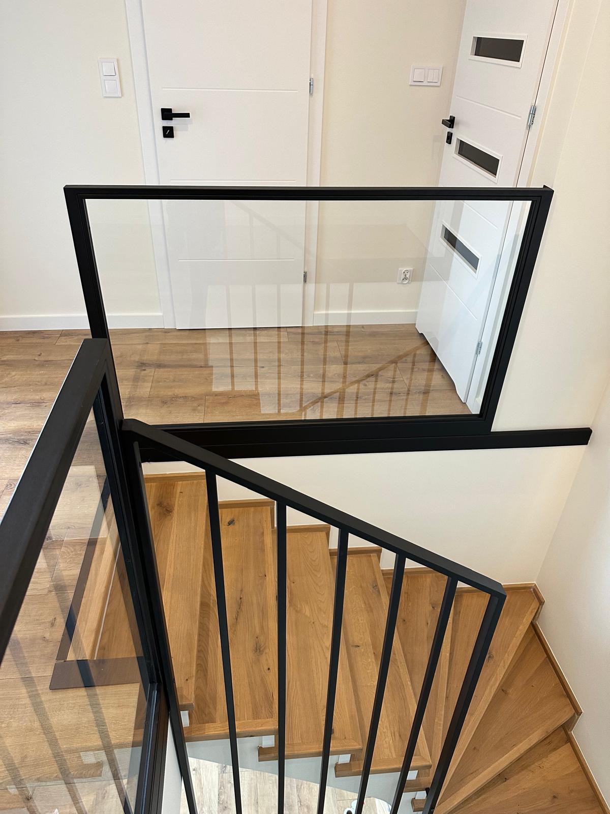 Nowoczesne schody drewniane z balustradą szklaną w czarnej ramie, widok z góry, dwie białe drzwi w tle, parkiet na podłodze. Minimalistyczny design.