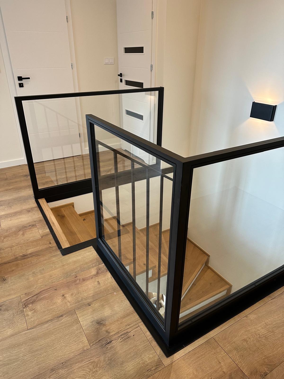 Nowoczesne schody z drewnianymi stopniami i balustradą ze szkła w czarnej ramie, oświetlone kinkietem. Minimalistyczny design.