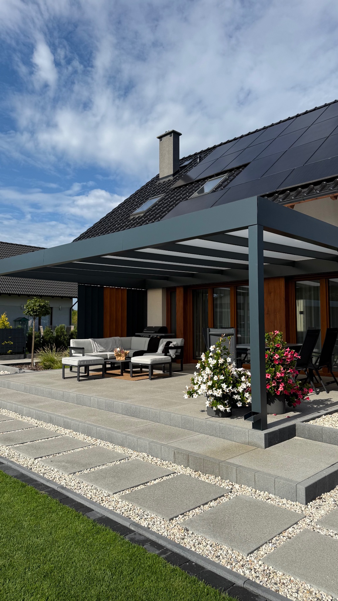 Nowoczesna pergola z szarych profili na tarasie z kostki, z widokiem na dom z panelami słonecznymi. Białe i różowe kwiaty w donicach dodają koloru. Słoneczny dzień.