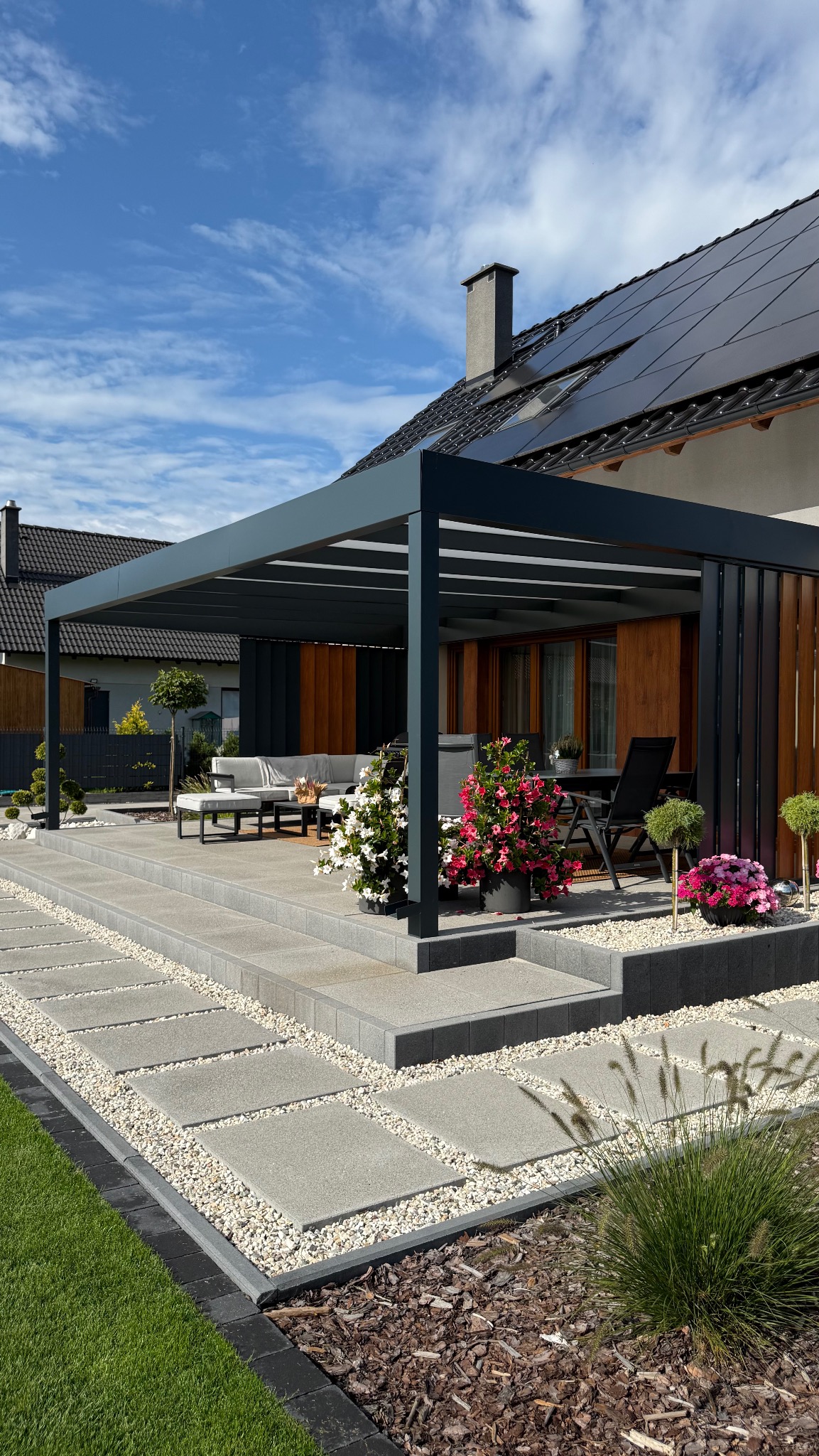 Nowoczesna, grafitowa pergola z lamelami na tarasie, z widokiem na meble ogrodowe i kwiaty w donicach. Tło stanowi dom z panelami słonecznymi i błękitne niebo.