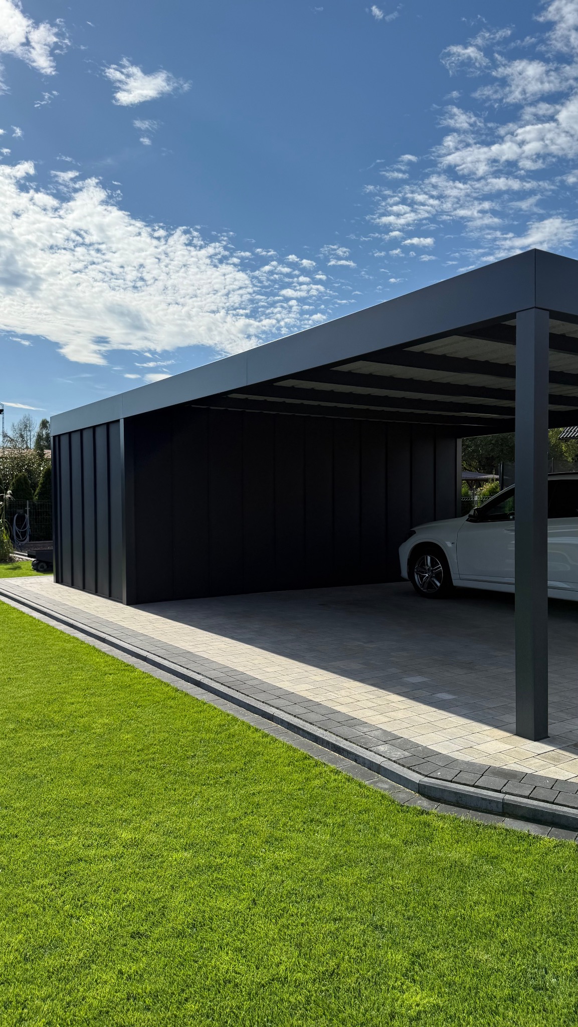 Nowoczesna, antracytowa wiata garażowa z częściowo zabudowanymi ścianami bocznymi, z białym samochodem w środku, na tle błękitnego nieba i zielonego trawnika.