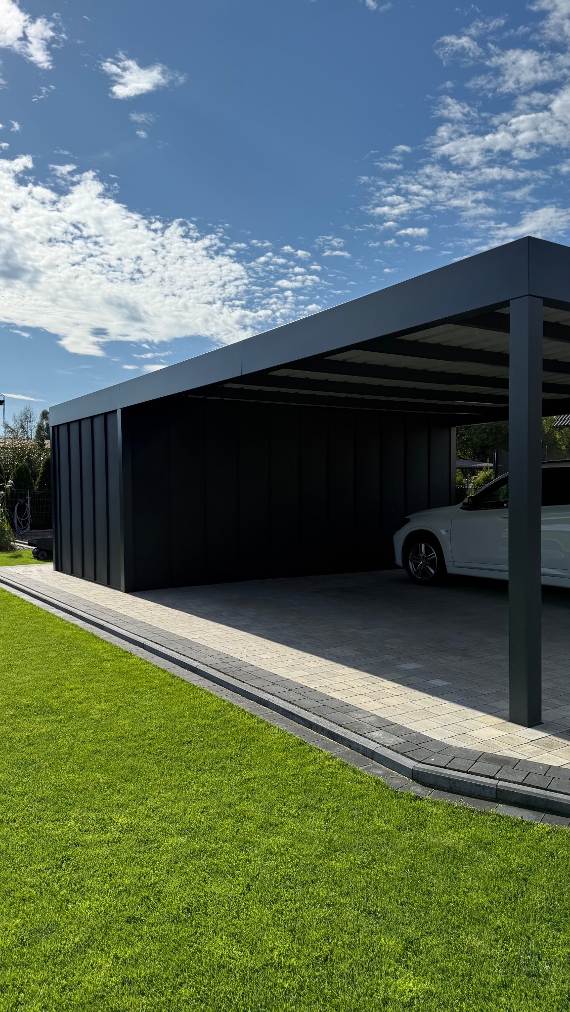 Nowoczesna, minimalistyczna pergola z metalu w antracytowym kolorze, przylegająca do budynku, z zaparkowanym samochodem w środku, na tle zielonego trawnika.