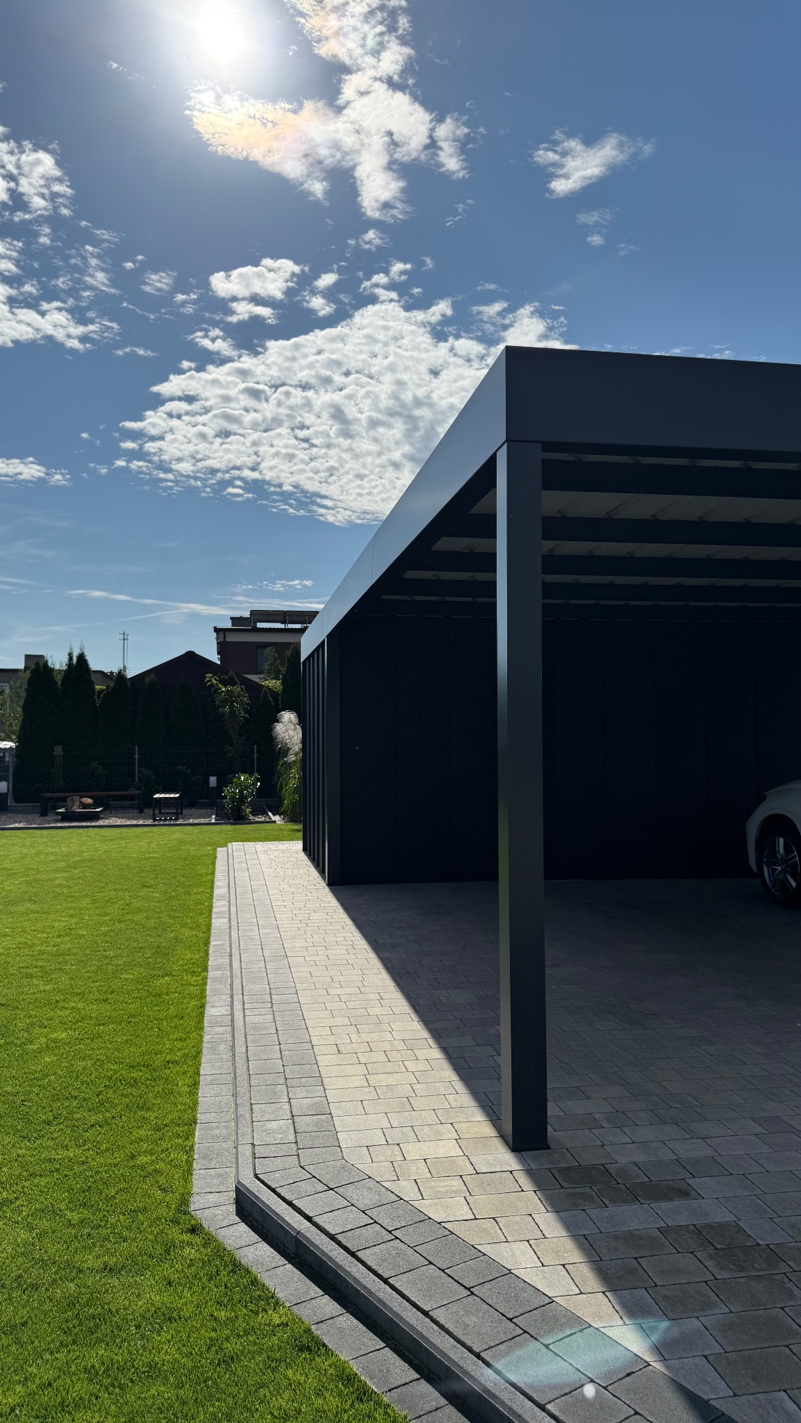 Nowoczesna, antracytowa pergola metalowa na posesji z równo przystrzyżonym trawnikiem i brukowanym podjazdem, w tle dom z ciemnym dachem i iglakami.