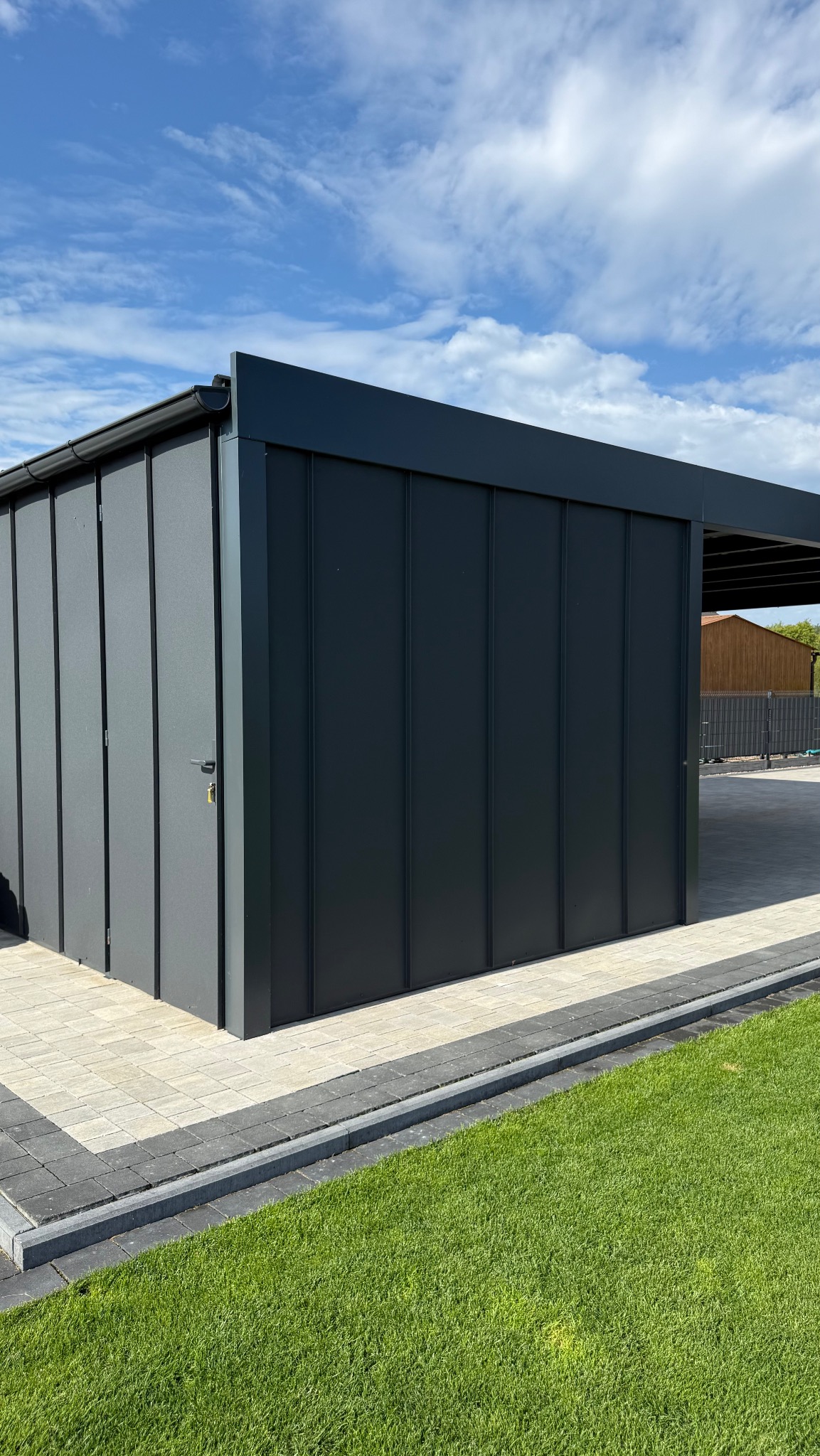 Nowoczesna, antracytowa pergola metalowa z zamkniętą częścią, widok z boku, na tle błękitnego nieba z chmurami i zielonego trawnika. Estetyczne wykończenie.