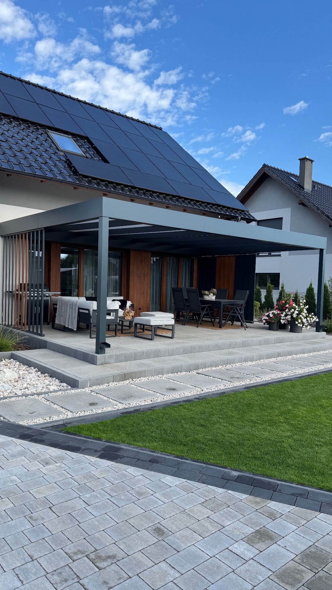Nowoczesna, szara pergola z elementami drewna, przylegająca do domu z panelami słonecznymi, otoczona zadbanym ogrodem. Na tarasie meble wypoczynkowe i stół.