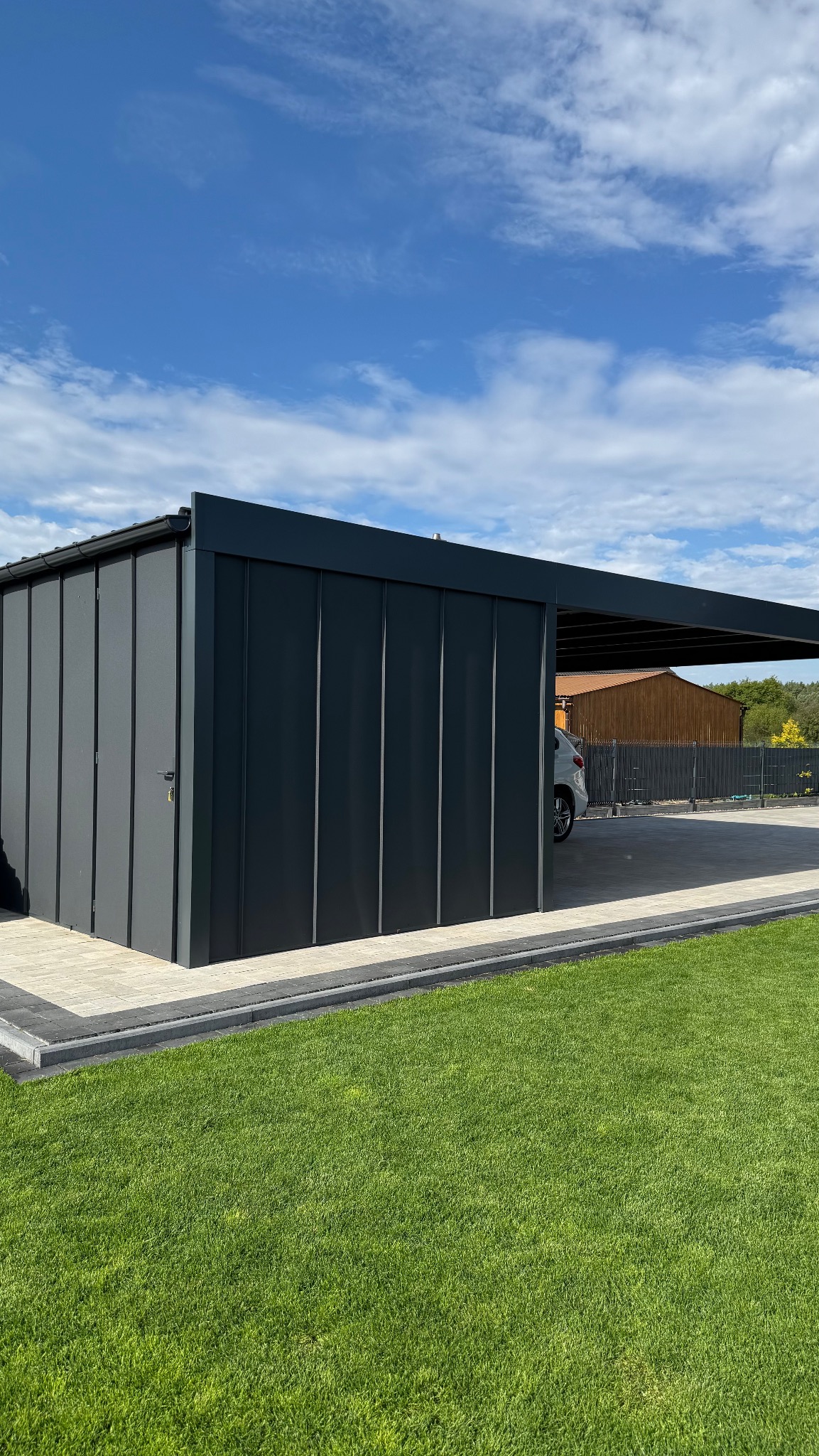 Nowoczesna, grafitowa wiata garażowa z zabudową, na tle błękitnego nieba i zielonego trawnika, z widocznym fragmentem zaparkowanego samochodu.