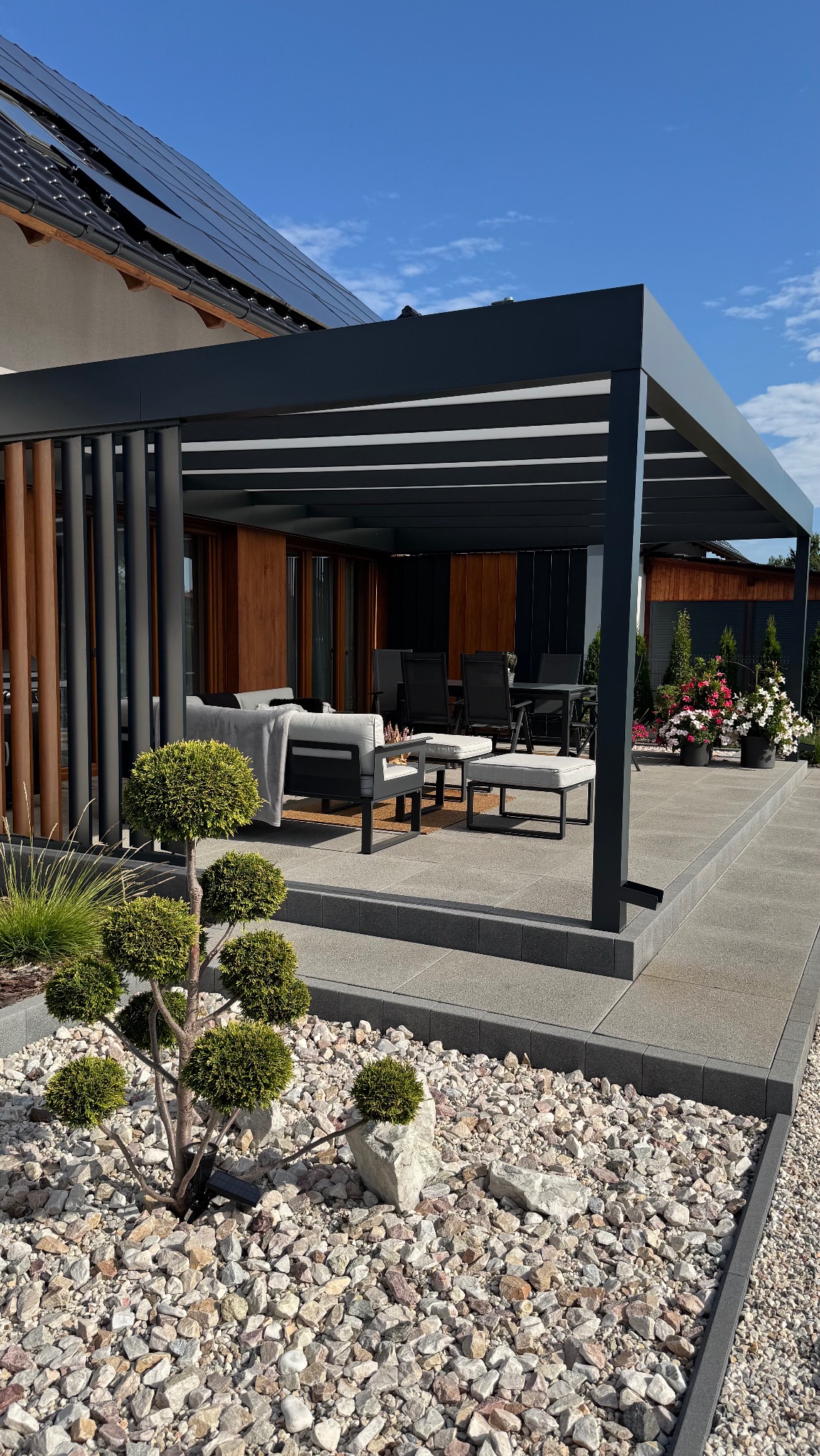 Nowoczesna, antracytowa pergola z lamelami, z meblami ogrodowymi, na tle domu z panelami słonecznymi i dekoracyjnym ogrodem z kamieniami i roślinami.