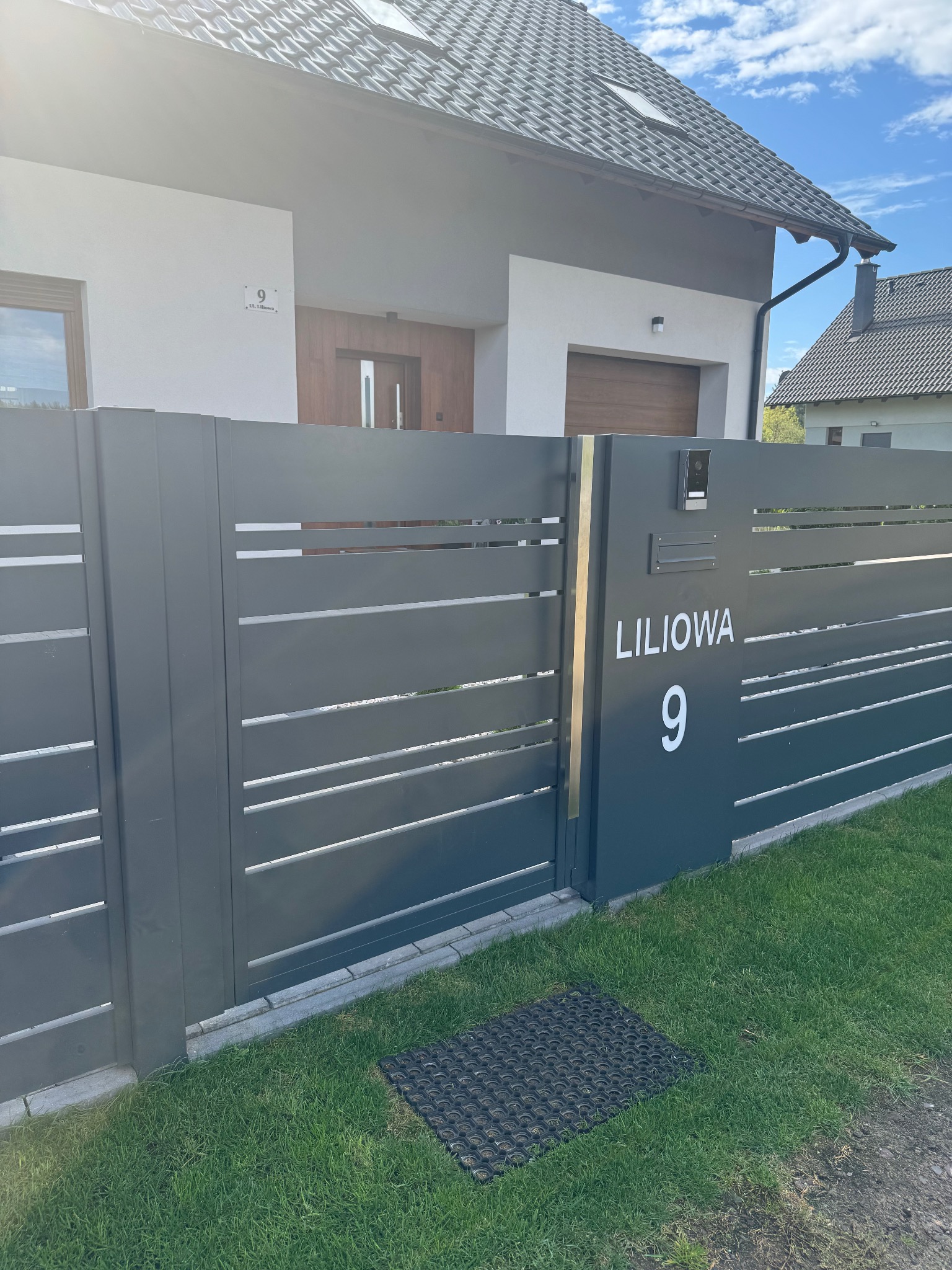 Nowoczesna, szara brama wjazdowa z poziomymi szczeblami, numerem domu 'Liliowa 9' i wideodomofonem, na tle domu z drewnianymi elementami.