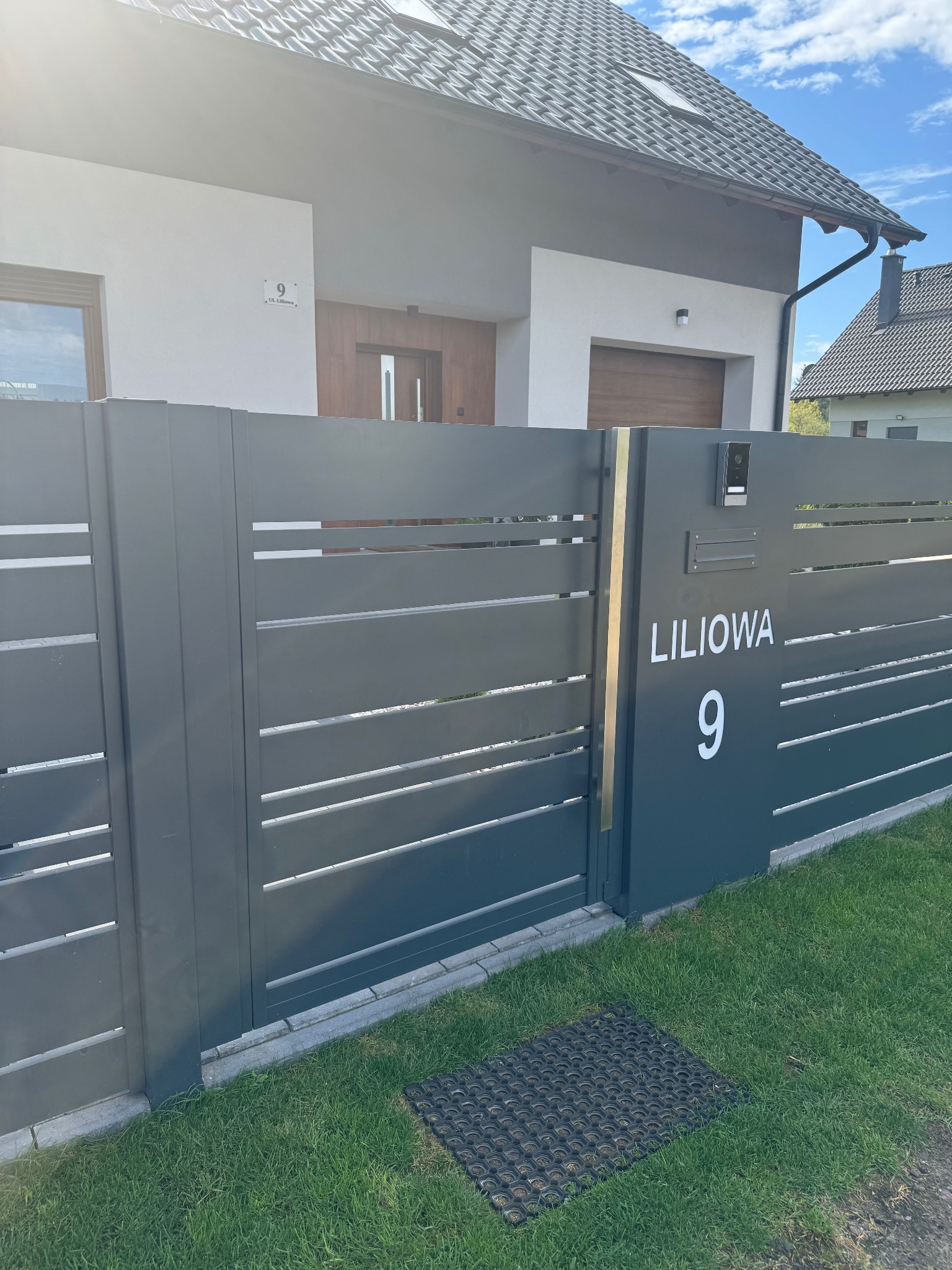 Nowoczesna brama wjazdowa w kolorze antracytowym z poziomymi panelami i numerem domu 'Liliowa 9', na tle jasnego domu z drewnianymi akcentami.