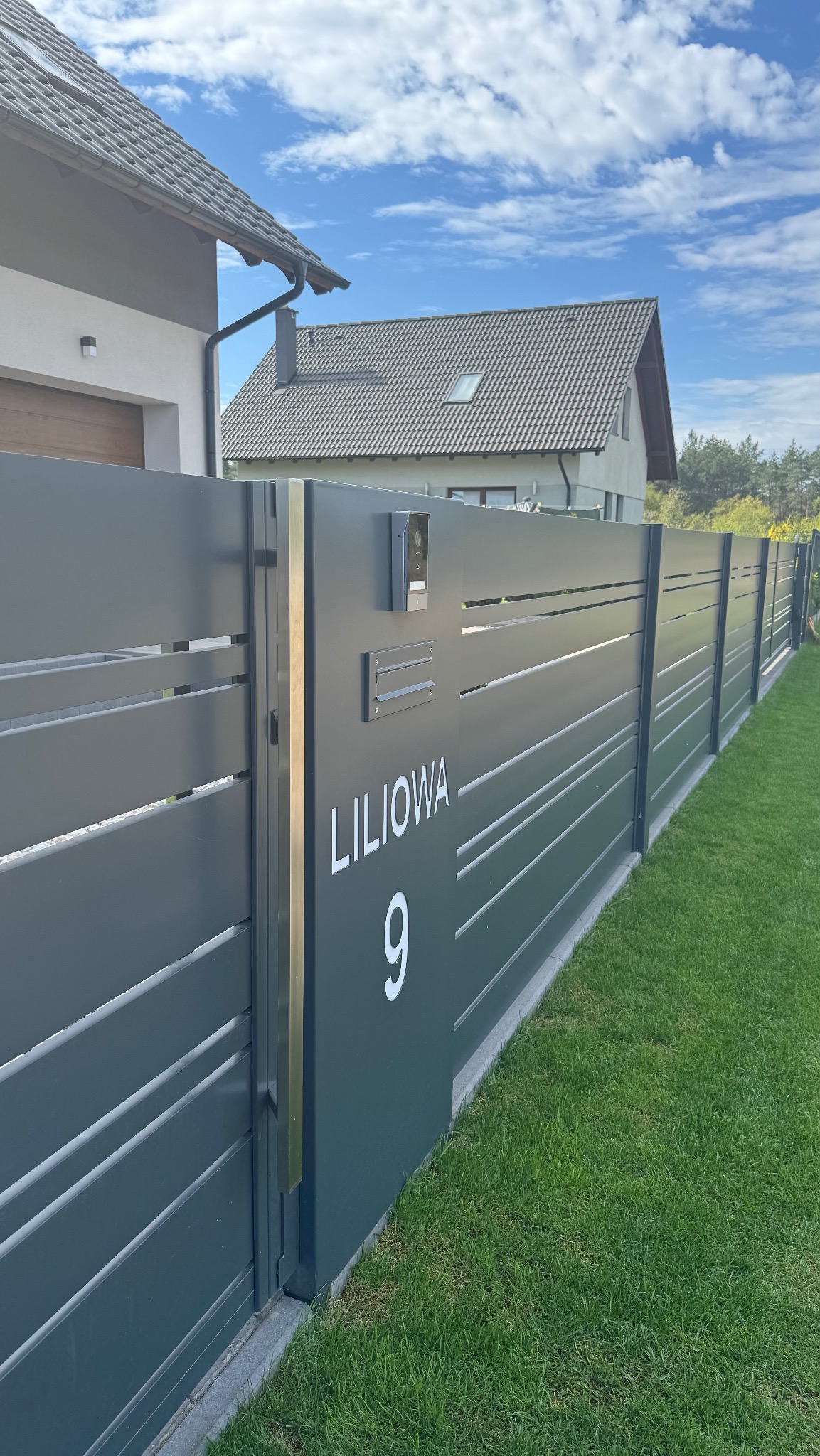 Nowoczesne, antracytowe ogrodzenie panelowe z adresem 'Liliowa 9', skrzynką pocztową i domofonem na tle zielonego trawnika i zabudowy jednorodzinnej.