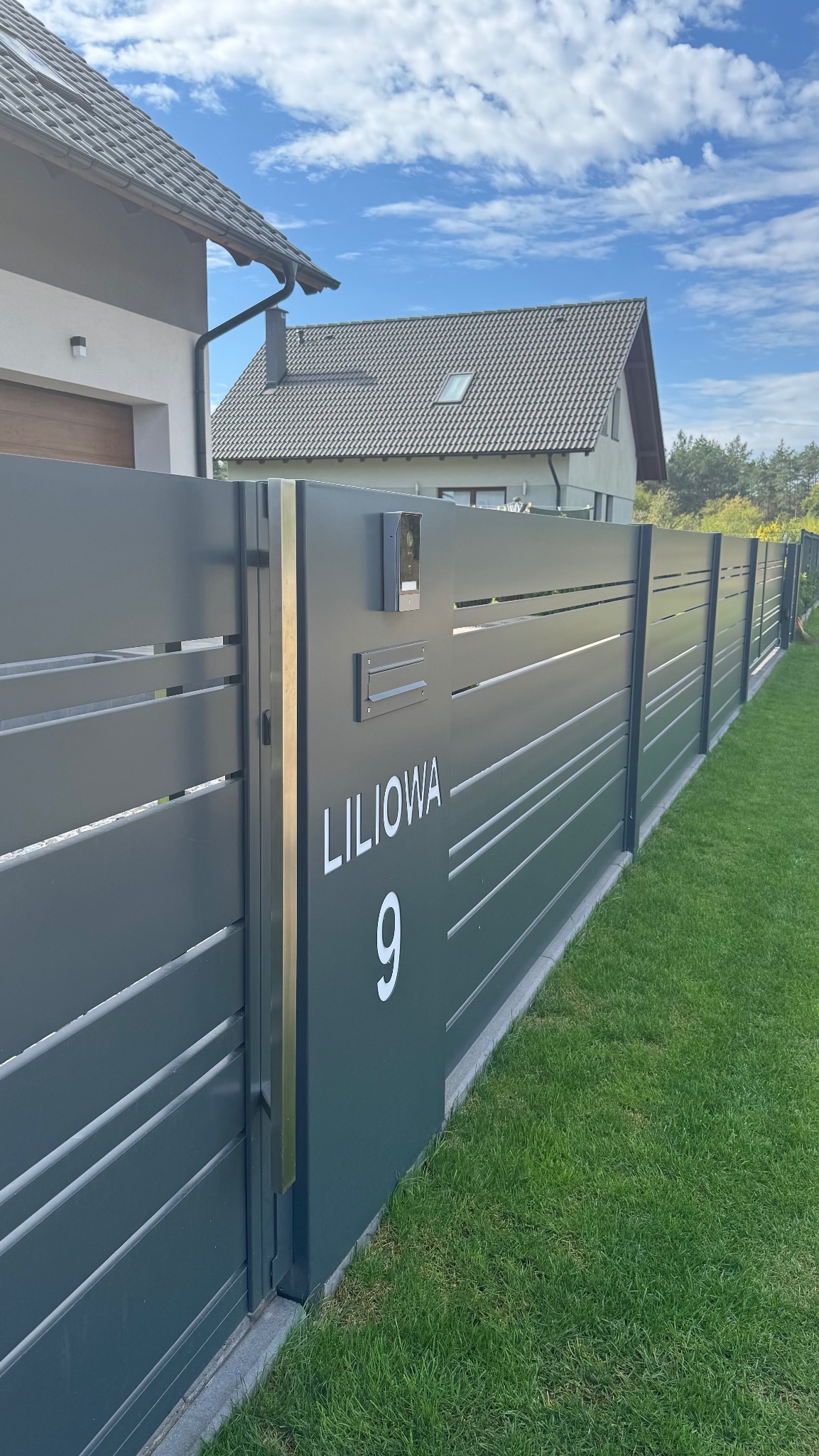 Nowoczesne, antracytowe ogrodzenie posesji z numerem domu 'Liliowa 9', z widoczną furtką i skrzynką na listy, na tle zielonego trawnika i domu.