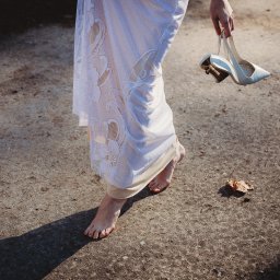 Historie Obrazkowe - Panna młoda boso w koronkowej sukni, trzymająca białe szpilki na tle betonowej nawierzchni z liściem.