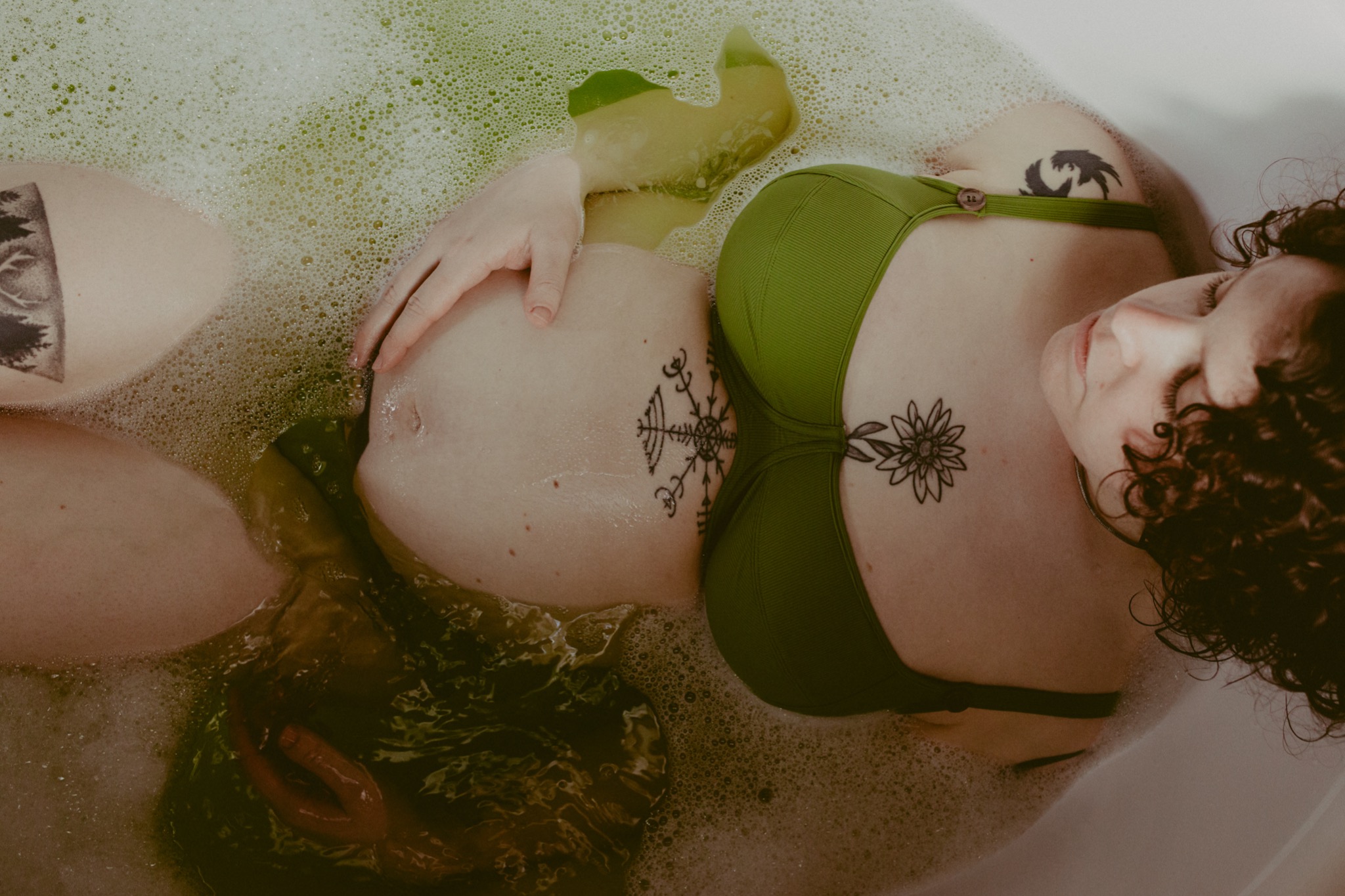 Kobieta w zielonym bikini relaksuje się w wannie wypełnionej wodą z bąbelkami i zielonymi liśćmi, eksponując ciążowy brzuszek z tatuażem.