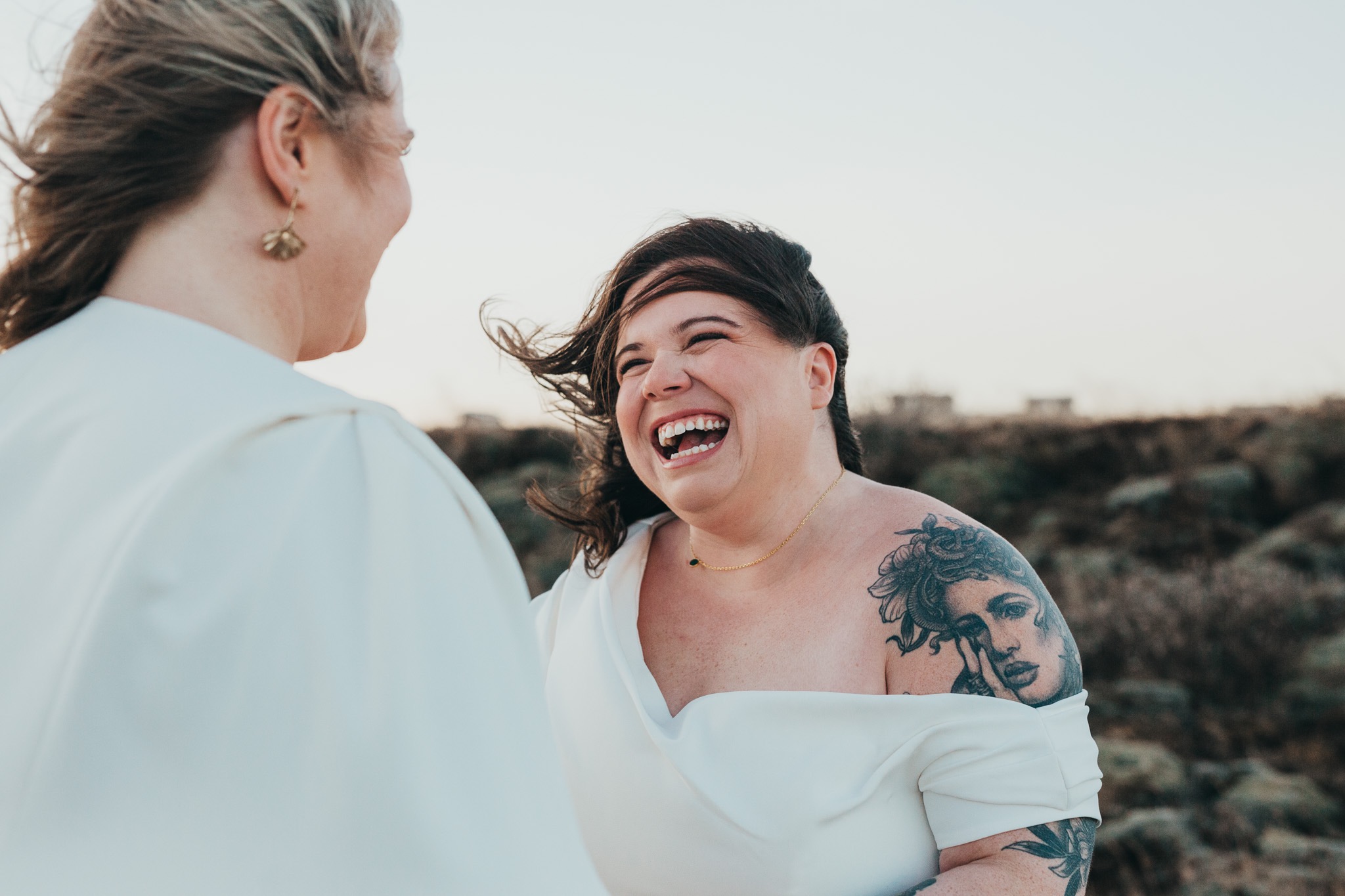 Dwie kobiety w jasnych sukienkach, jedna z tatuażami na ramionach, śmieją się do siebie na tle krajobrazu z niską roślinnością.