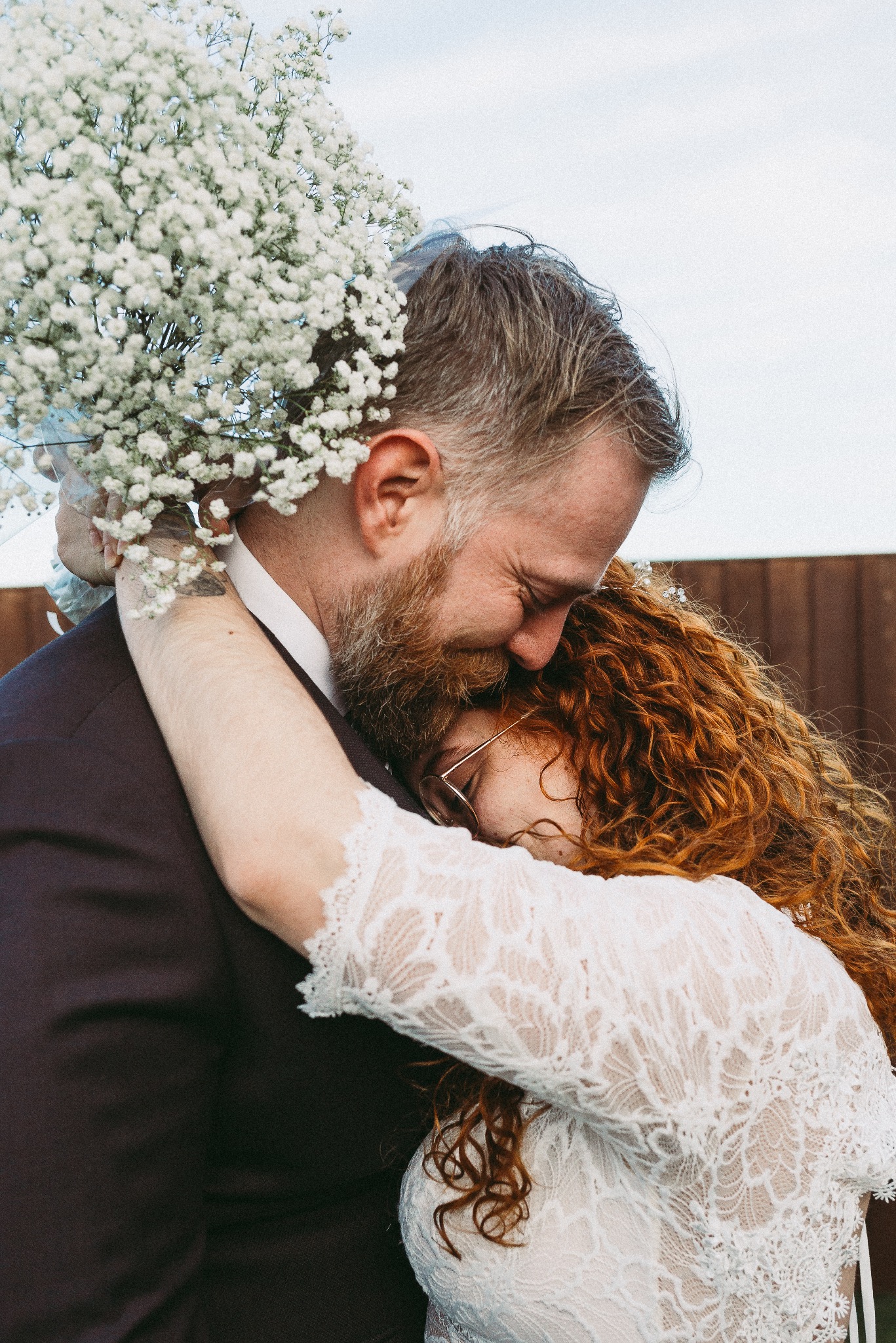 Para młoda w Białymstoku w czułym uścisku. Panna młoda z rudymi włosami i okularami, ubrana w koronkową suknię, opiera głowę o ramię pana młodego w ciemnym garniturze. W tle bukiet z białych kwiatów.