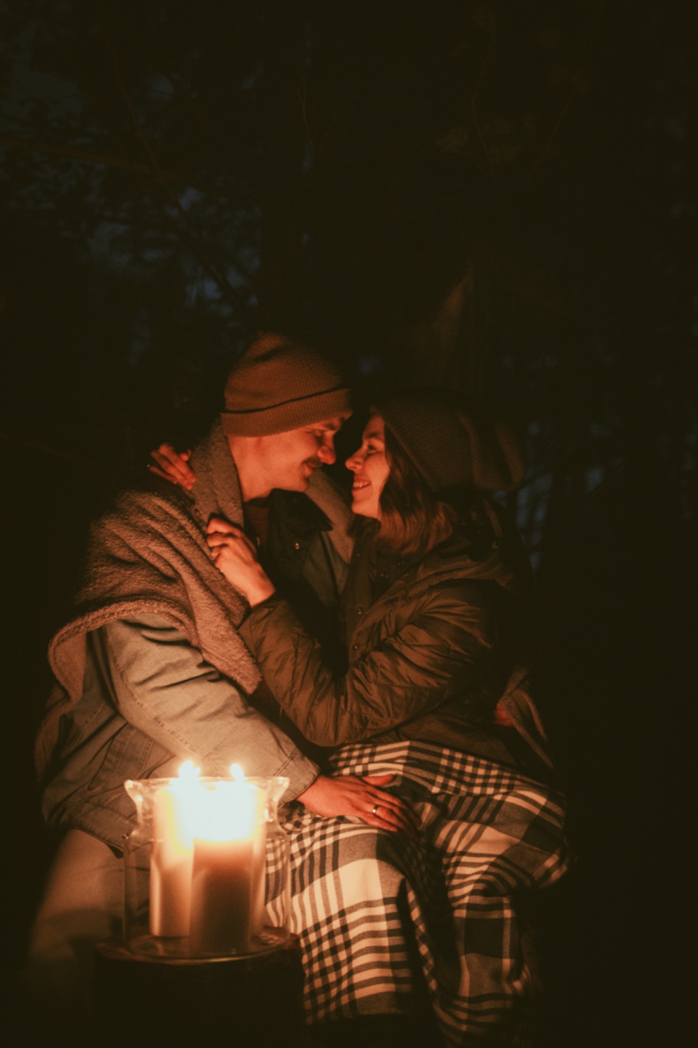 Para w czapkach i kurtkach przytulona w ciemności, oświetlona ciepłym blaskiem świec w szklanym lampionie, okryta pledem w kratę.