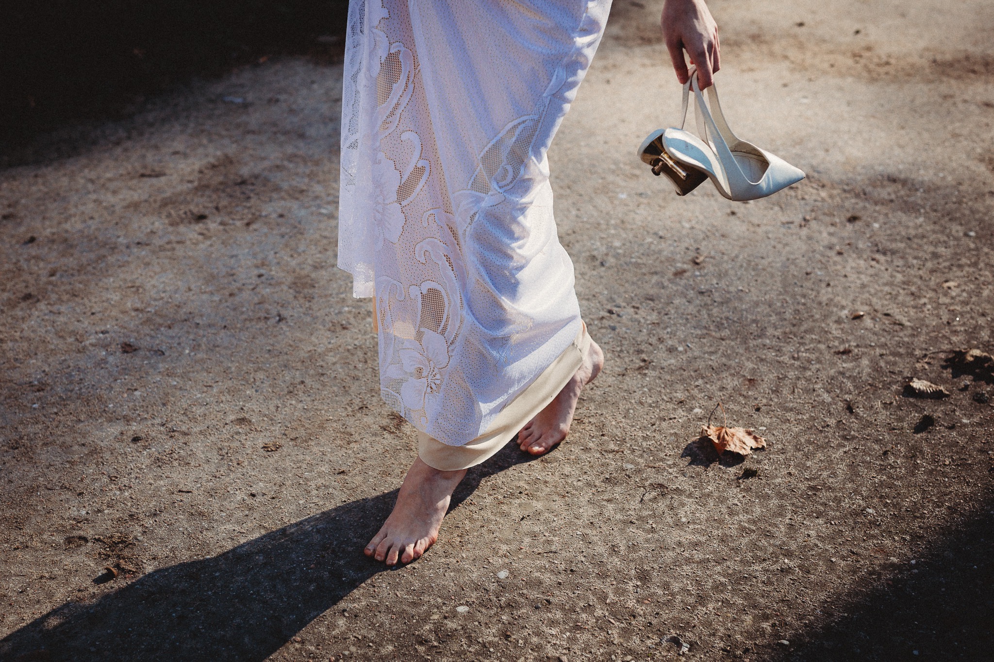 Panna młoda boso w koronkowej sukni, trzymająca białe szpilki na tle betonowej nawierzchni z liściem.