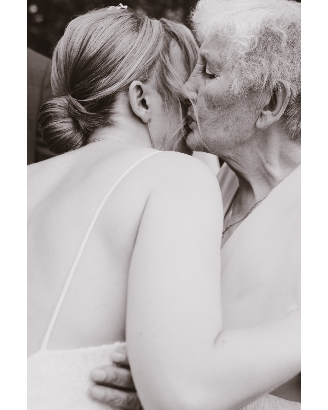 Czarno-białe zbliżenie panny młodej w sukni na ramiączkach obejmującej starszą kobietę, prawdopodobnie babcię, która ją całuje w policzek, z widocznym upięciem włosów panny młodej i delikatną...