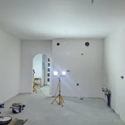 Wnętrze w trakcie prac w Warszawie: ściany przygotowane do malowania, widoczne ceglane elementy dekoracyjne, lampy budowlane oświetlają przestrzeń, podłoga zabezpieczona. Częściowo widoczne...