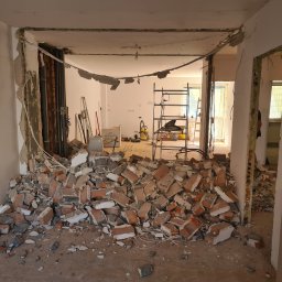 VADYM NIKOLAIEV UBR - Demontaż ściany działowej z cegły w trakcie remontu mieszkania. Widoczne instalacje, gruz i rusztowanie w tle. Ujęcie z perspektywy wnętrza.