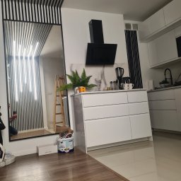 VADYM NIKOLAIEV UBR - Nowoczesna kuchnia z białymi meblami, czarnym okapem i lustrem z czarnymi listwami. Na podłodze panele imitujące drewno. Widoczny fragment drabiny malarskiej.