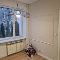 VADYM NIKOLAIEV UBR - Jasne wnętrze po remoncie: kremowe ściany z dekoracyjną ramą, nowoczesna lampa, okno z widokiem na budynek. Na podłodze puszka z farbą i karton.