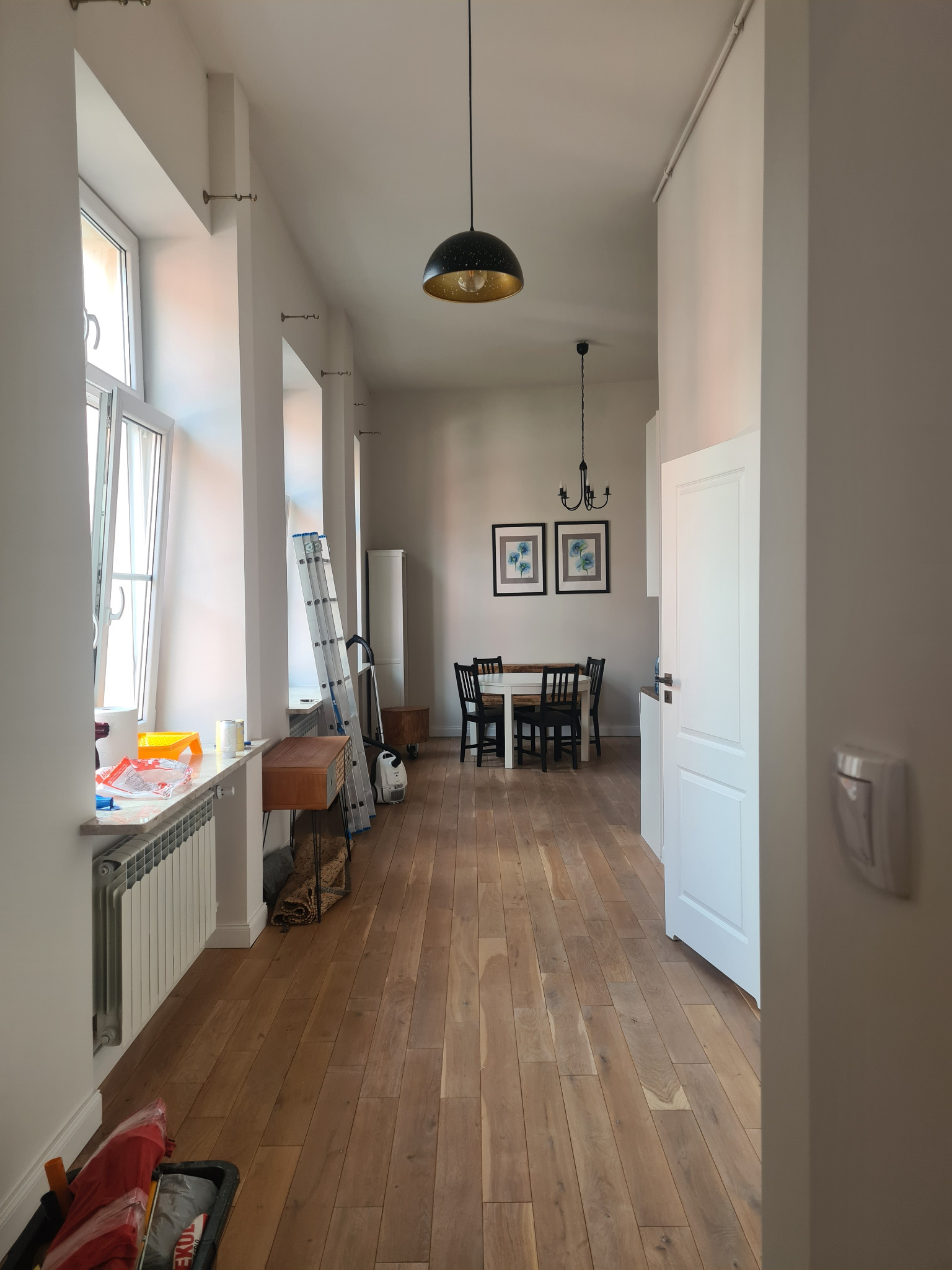 Wnętrze mieszkania w trakcie remontu: białe ściany, drewniana podłoga, drabina oparta o ścianę, akcesoria malarskie na parapecie, widok na jadalnię z białym stołem i czarnymi krzesłami.