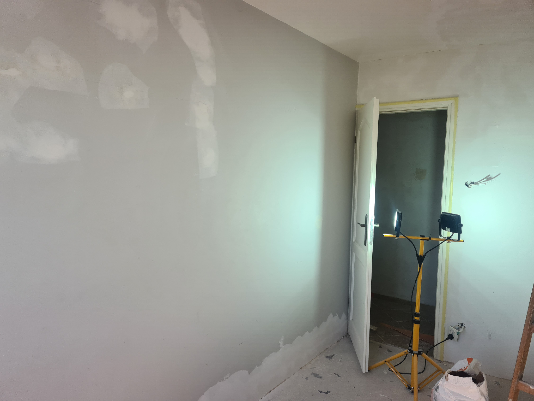 Pokój w trakcie przygotowania do malowania: ściany z miejscowymi naprawami i szpachlowaniem, oświetlone halogenem na statywie, otwarte drzwi z taśmą malarską na framudze.