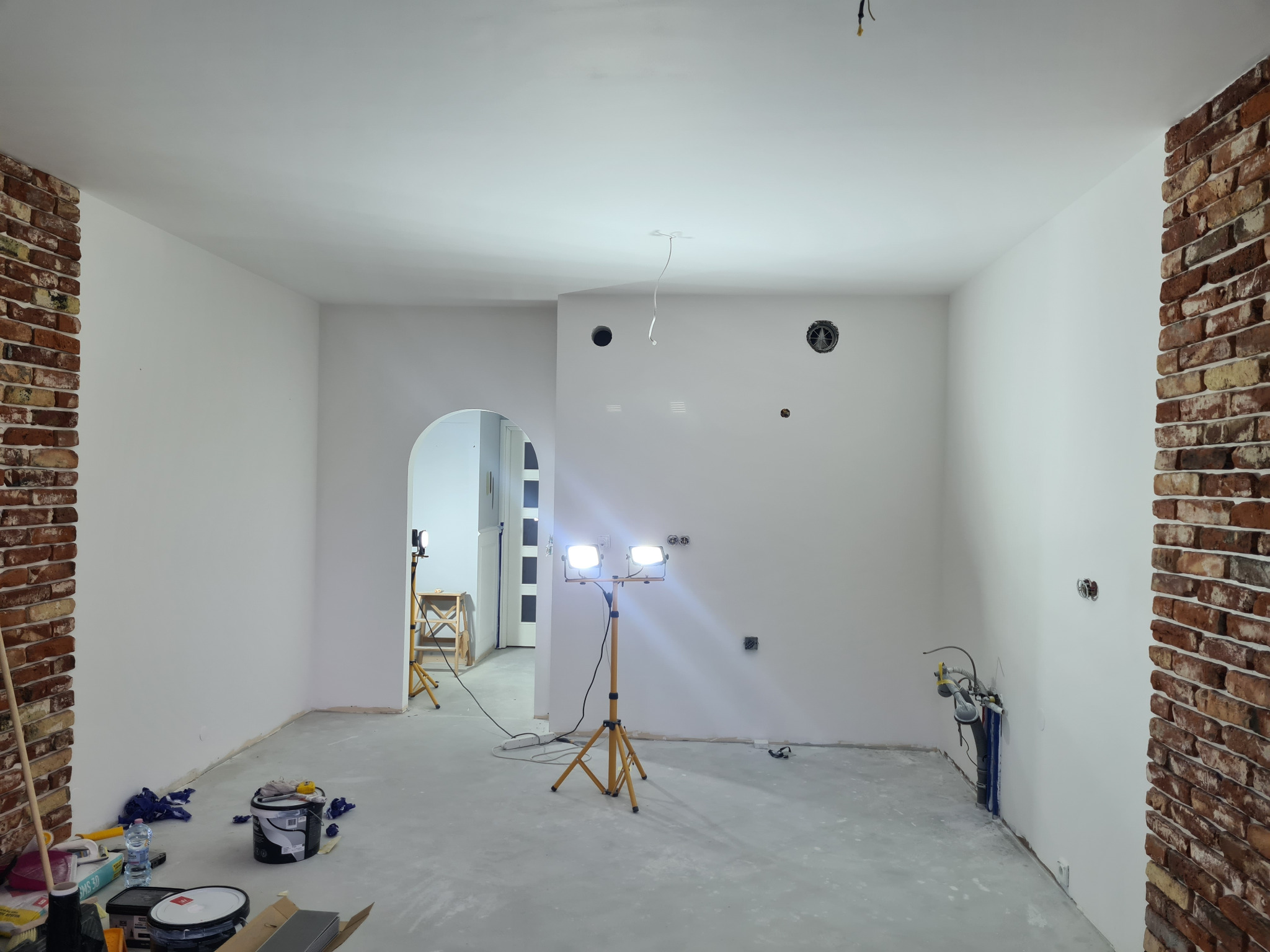 Wnętrze w trakcie prac w Warszawie: ściany przygotowane do malowania, widoczne ceglane elementy dekoracyjne, lampy budowlane oświetlają przestrzeń, podłoga zabezpieczona. Częściowo widoczne...