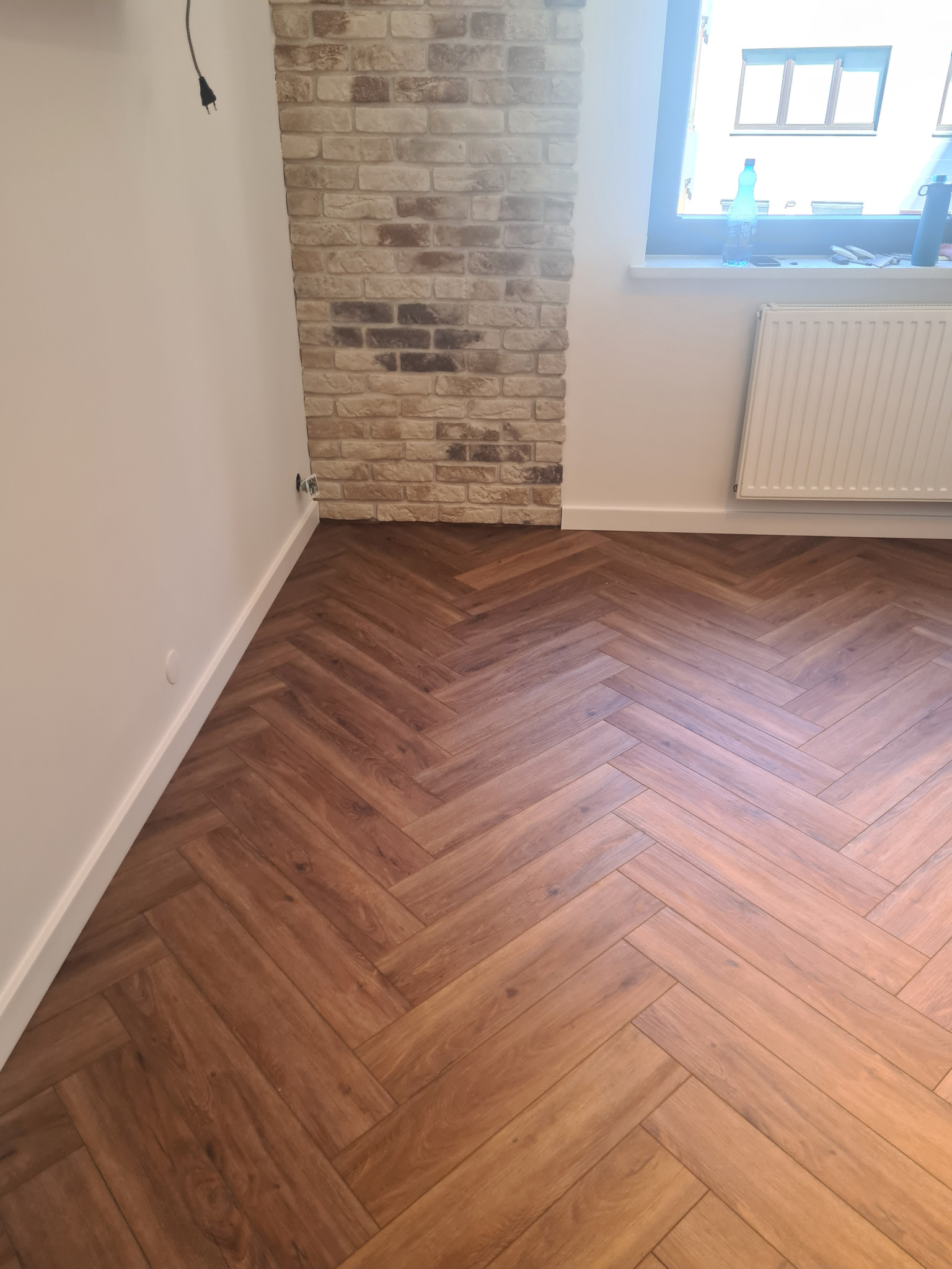 Ułożone panele podłogowe w jodełkę, widok na fragment ściany z dekoracyjną cegłą, białą ścianę i okno z grzejnikiem.