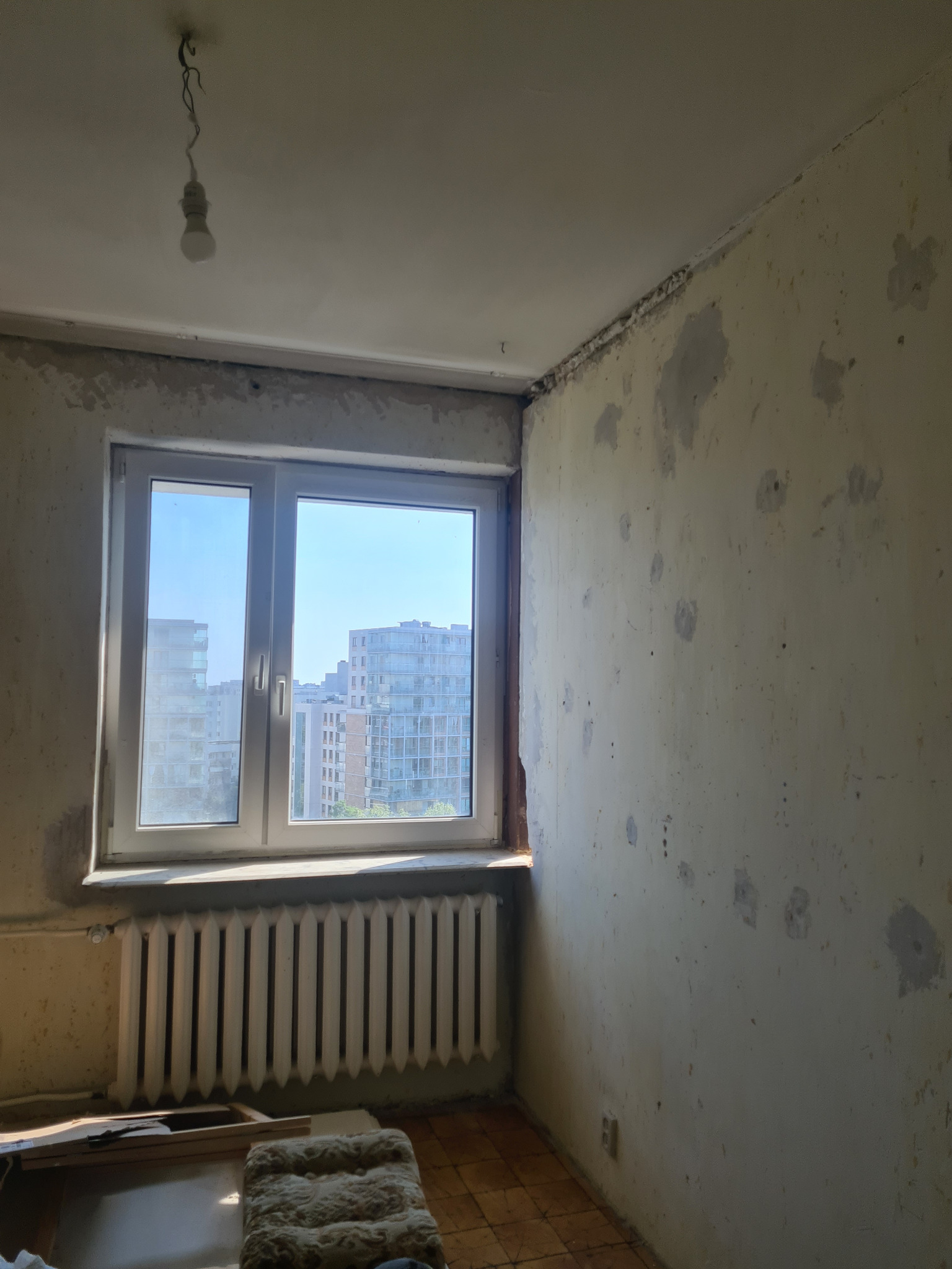 Pokój przed remontem: ściany z widocznymi śladami po naprawach, grzejnik pod oknem z widokiem na nowoczesne budynki miejskie, surowa żarówka zwisająca z sufitu.