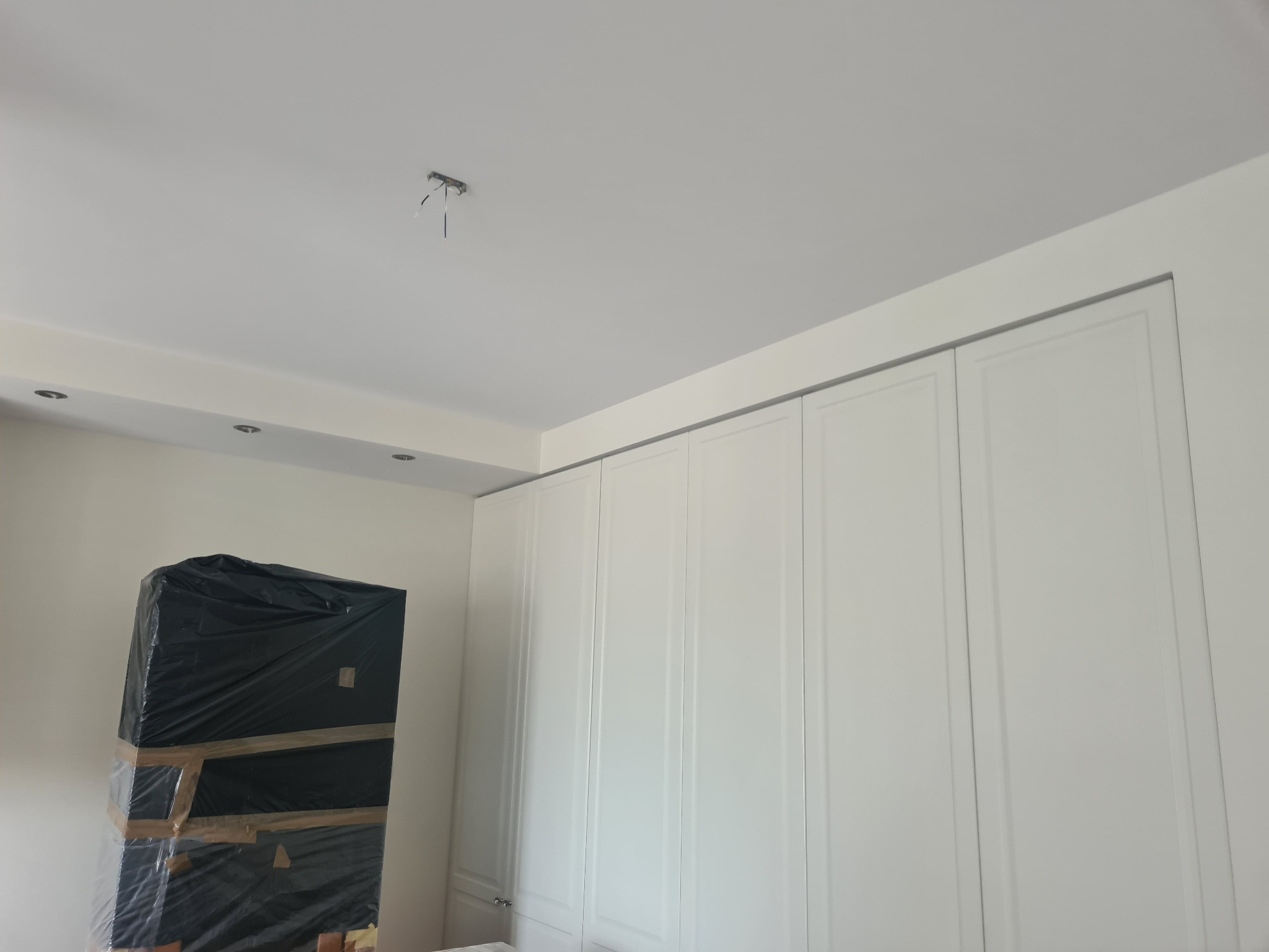 Nowo zamontowana biała szafa wnękowa z widocznym, niezabezpieczonym przewodem oświetleniowym na suficie oraz meblem zabezpieczonym folią malarską.