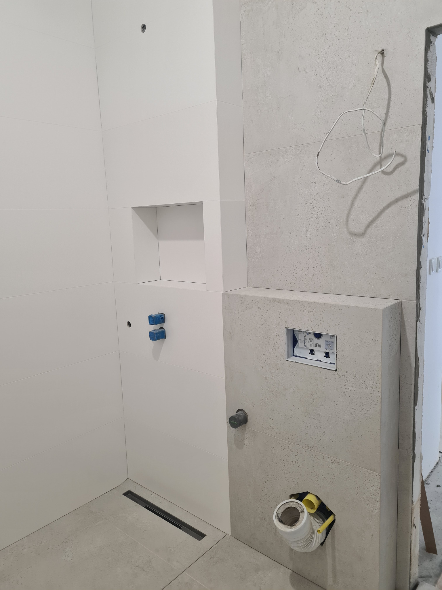 Wnętrze łazienki w trakcie remontu, widoczne białe i szare płytki, zabudowa geberitu z instalacją, odpływ liniowy w podłodze, surowe kable wystające ze ściany.