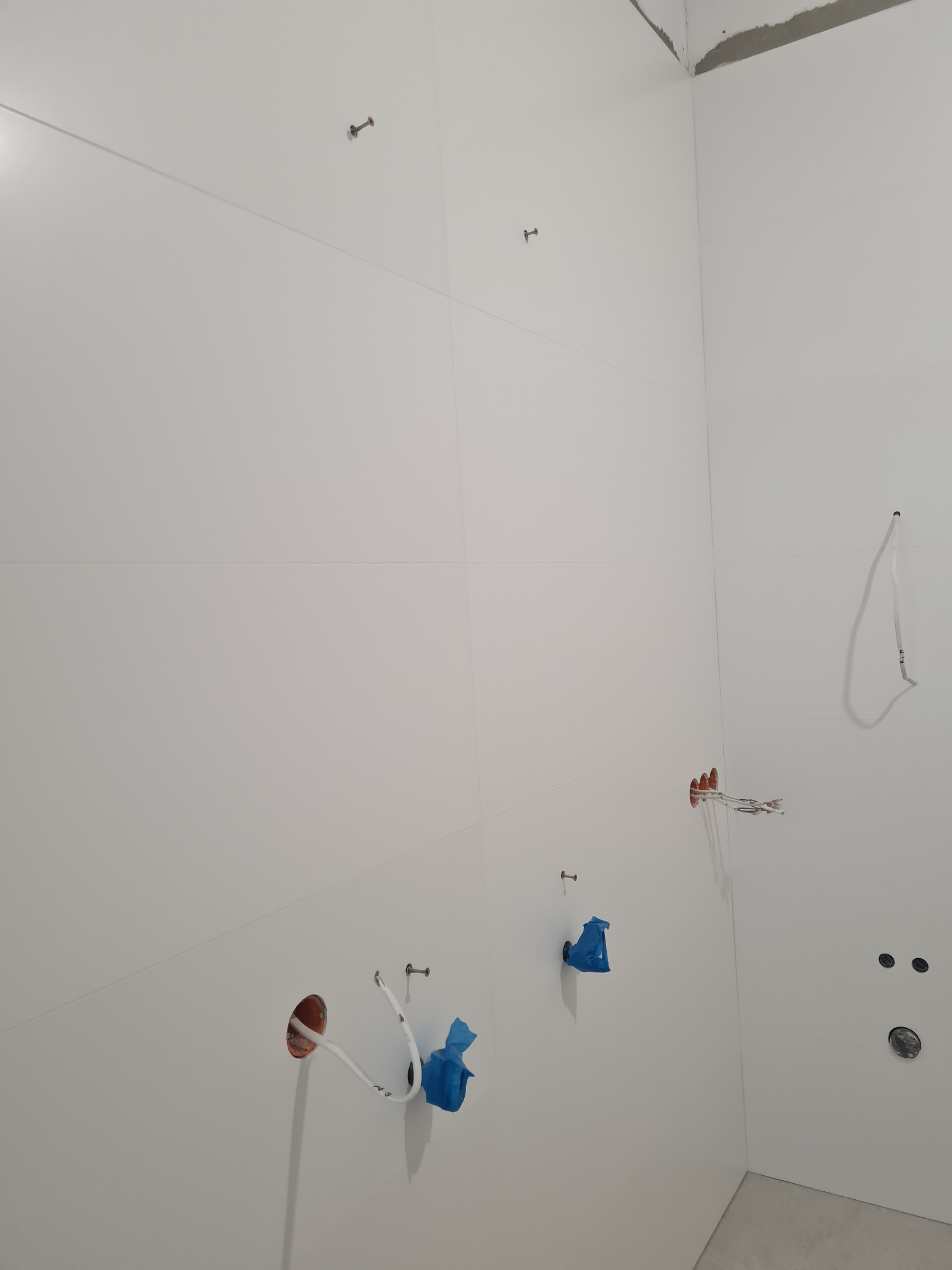 Wnętrze w trakcie prac wykończeniowych, ściany pokryte białymi płytami, widoczne otwory instalacyjne z wystającymi przewodami zabezpieczonymi niebieską taśmą, oraz metalowe kołki mocujące.
