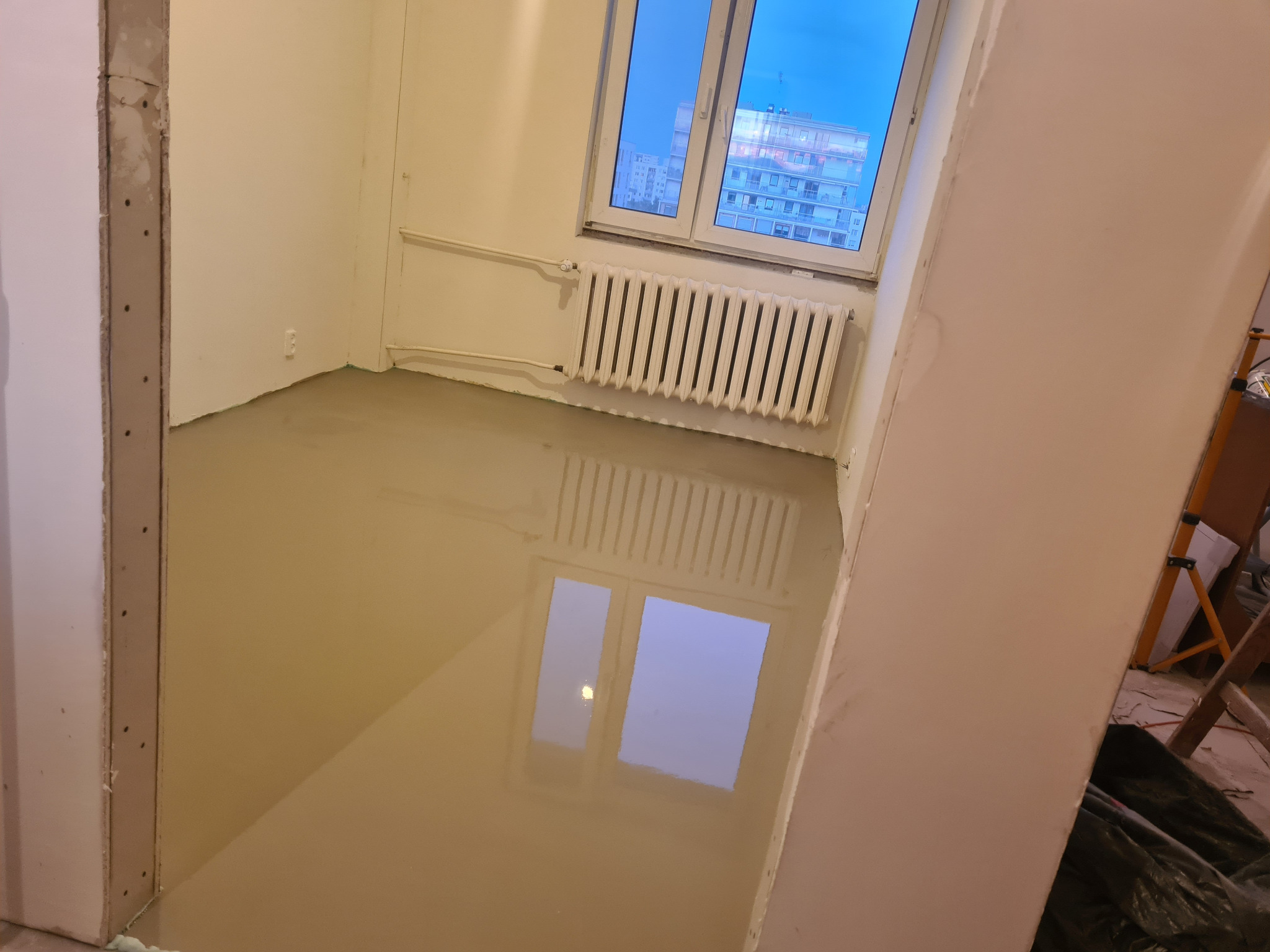 Wylewka samopoziomująca w pokoju z grzejnikiem i oknem, widoczne przygotowane ościeżnice drzwiowe, odbicie okna w mokrej powierzchni.