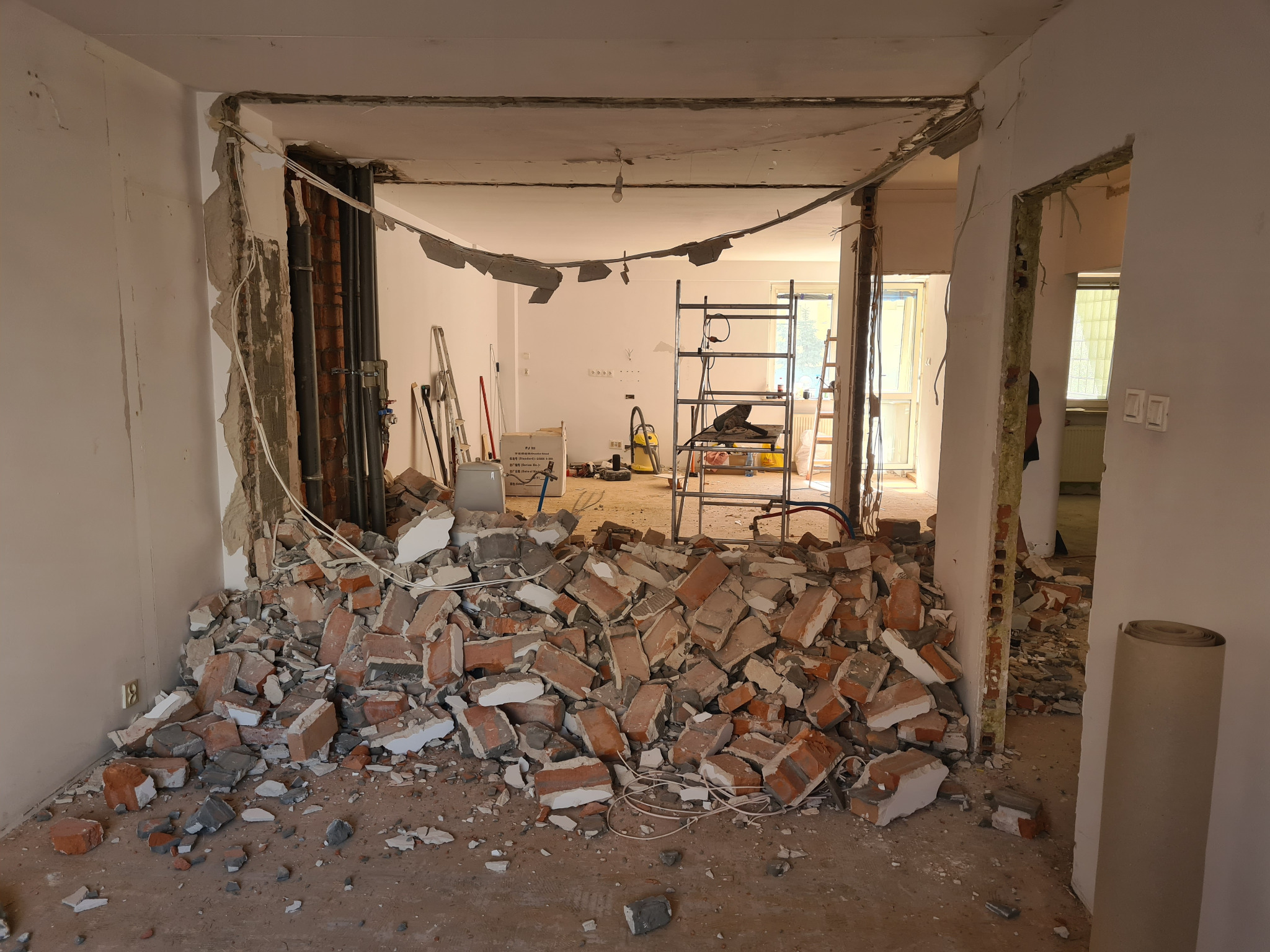 Demontaż ściany działowej z cegły w trakcie remontu mieszkania. Widoczne instalacje, gruz i rusztowanie w tle. Ujęcie z perspektywy wnętrza.
