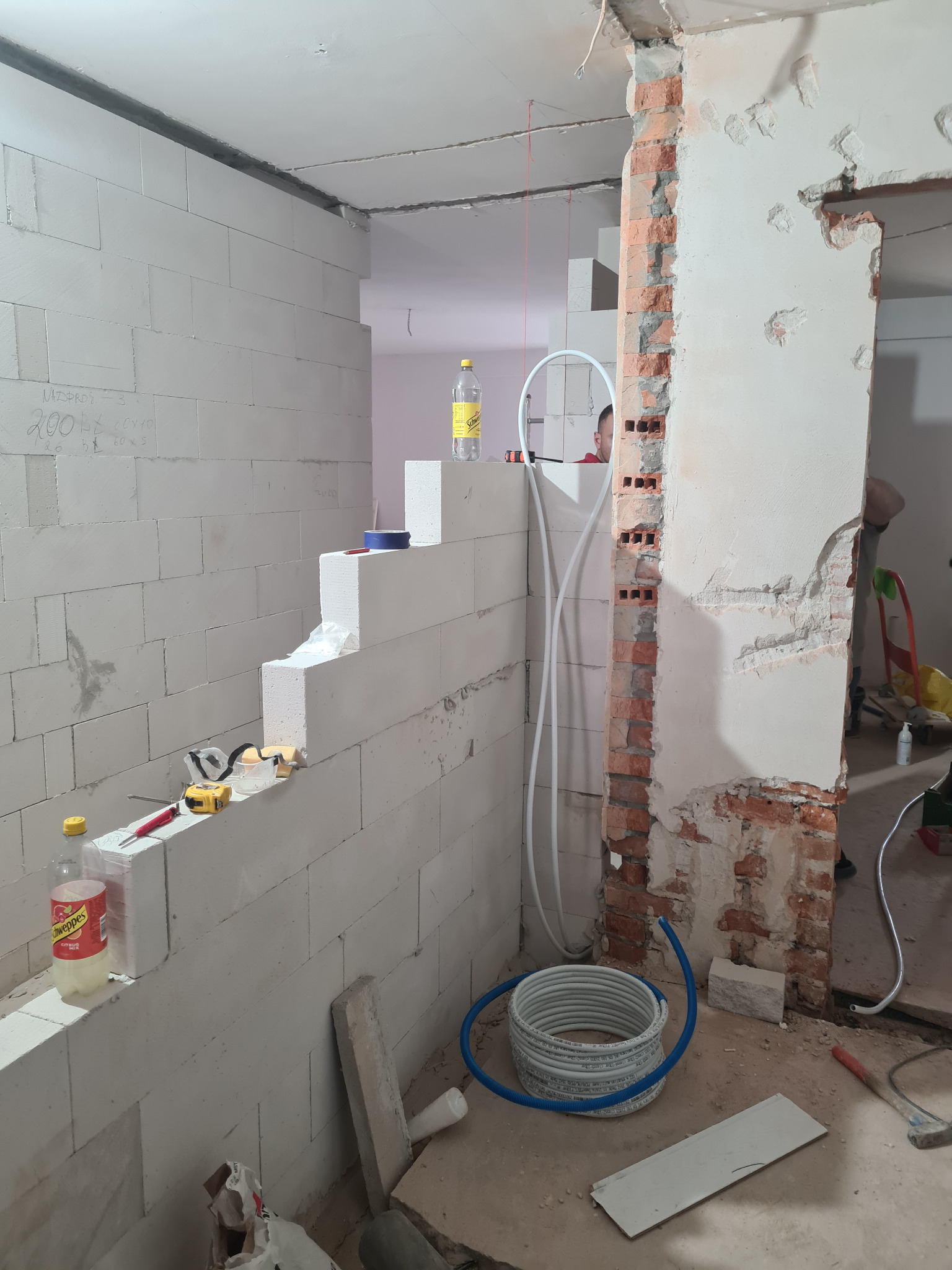 Wnętrze w trakcie remontu: ściana z białych bloczków, instalacja wodna, fragment ściany z cegły. Widoczne narzędzia i materiały budowlane. Prace murarskie i instalacyjne w toku.