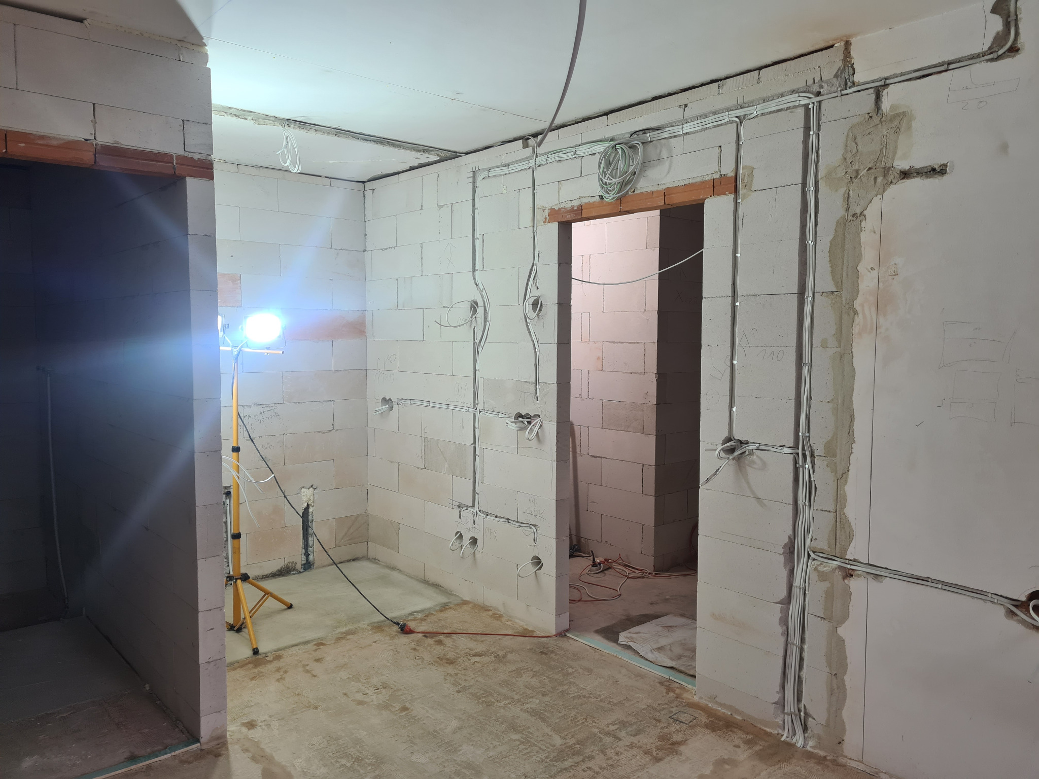 Wnętrze w trakcie prac instalacyjnych: widoczne przewody elektryczne prowadzone po ścianach z betonu komórkowego, surowy beton na podłodze, lampa budowlana.