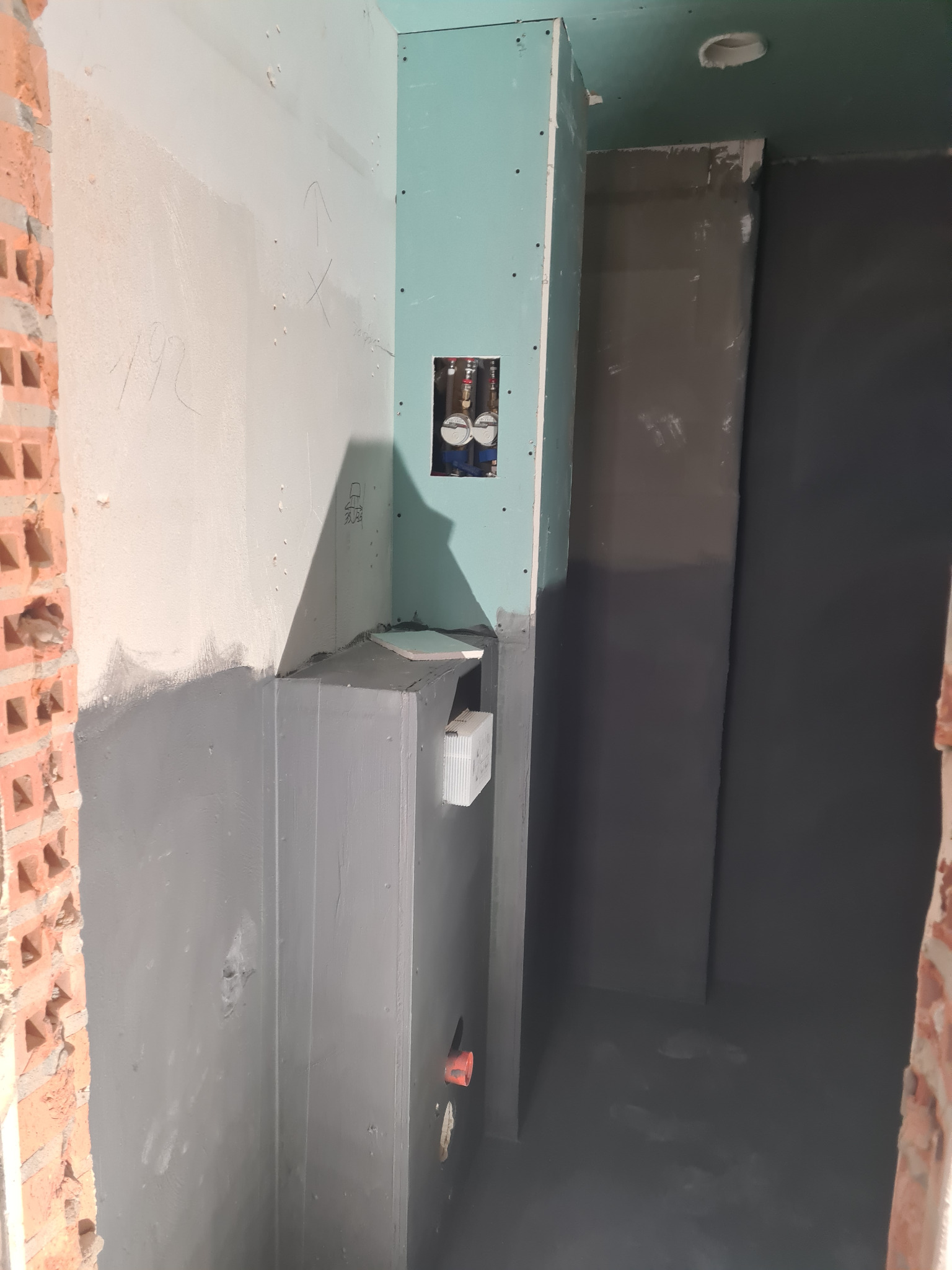 Wnętrze łazienki w trakcie remontu: ściany z płyt karton-gips, fragment instalacji wodnej, hydroizolacja w kolorze szarym, surowa cegła. Widok z dołu, oświetlenie naturalne.