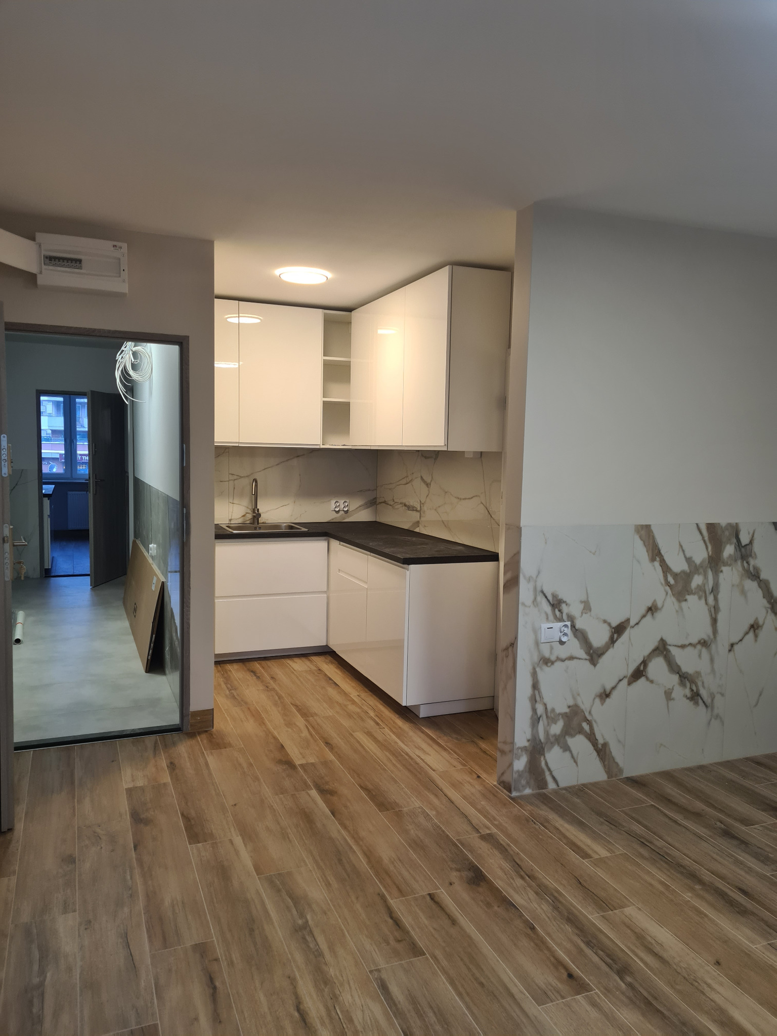 Nowoczesna kuchnia z białymi szafkami i marmurowym blatem, drewnopodobne panele podłogowe, widok na sąsiednie pomieszczenie w trakcie prac wykończeniowych.