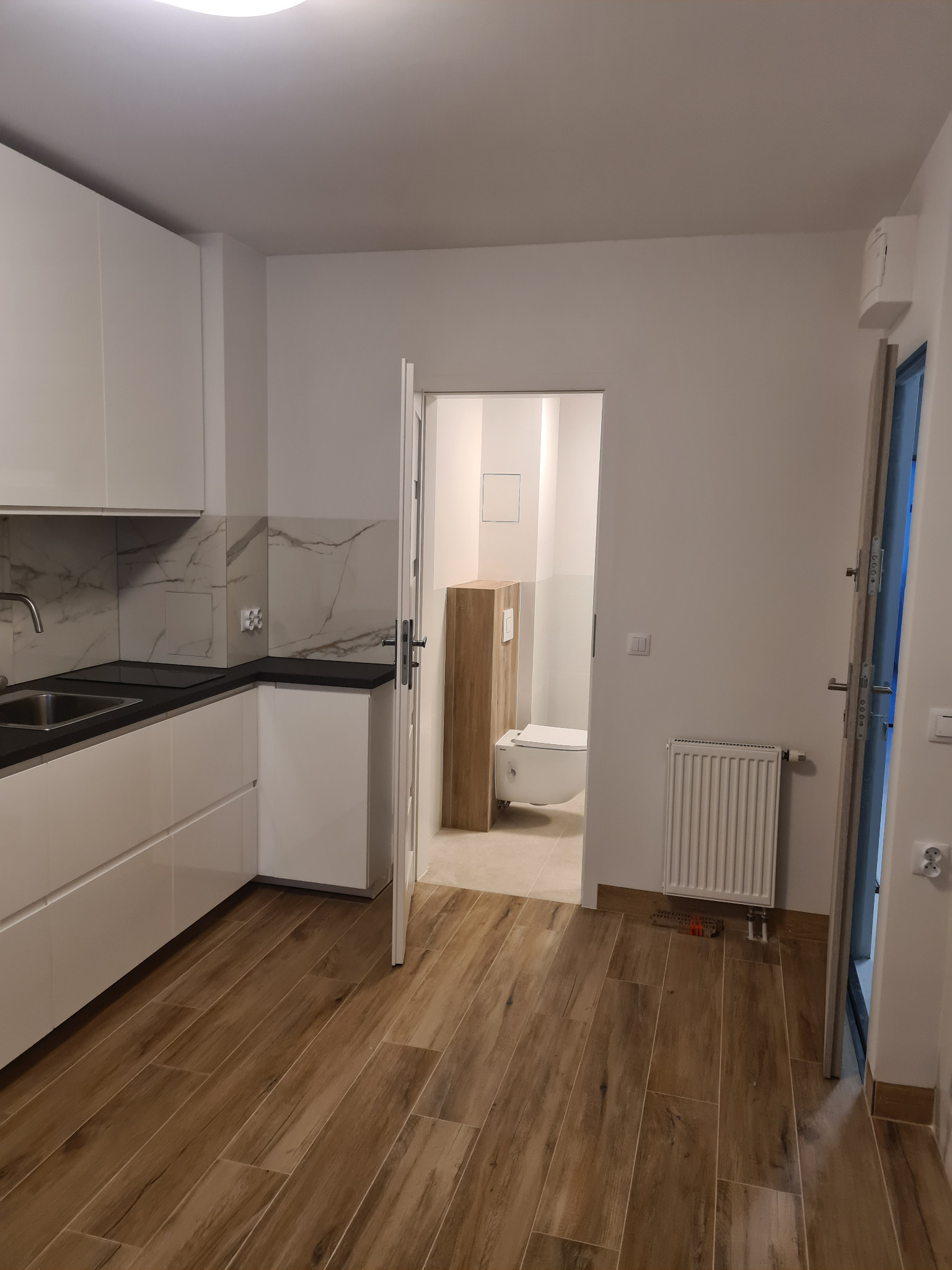 Nowoczesna kuchnia z białymi szafkami i marmurowym backsplashiem, widok na łazienkę z drewnianymi akcentami i brązowymi panelami podłogowymi. Minimalistyczny design.