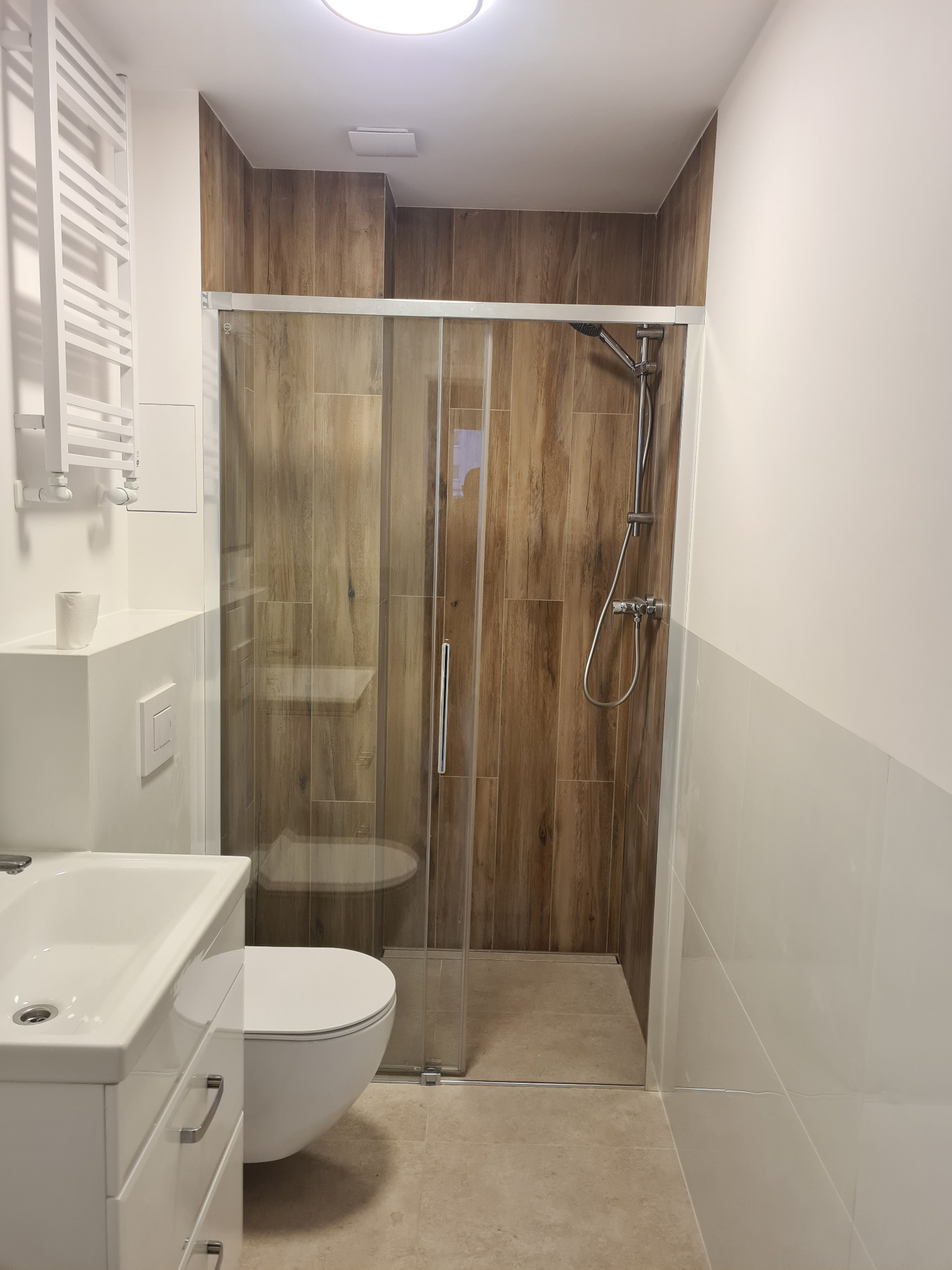 Nowoczesna łazienka z kabiną prysznicową typu walk-in, wykończona płytkami imitującymi drewno i białymi płytkami. Widoczna umywalka z szafką i toaleta.