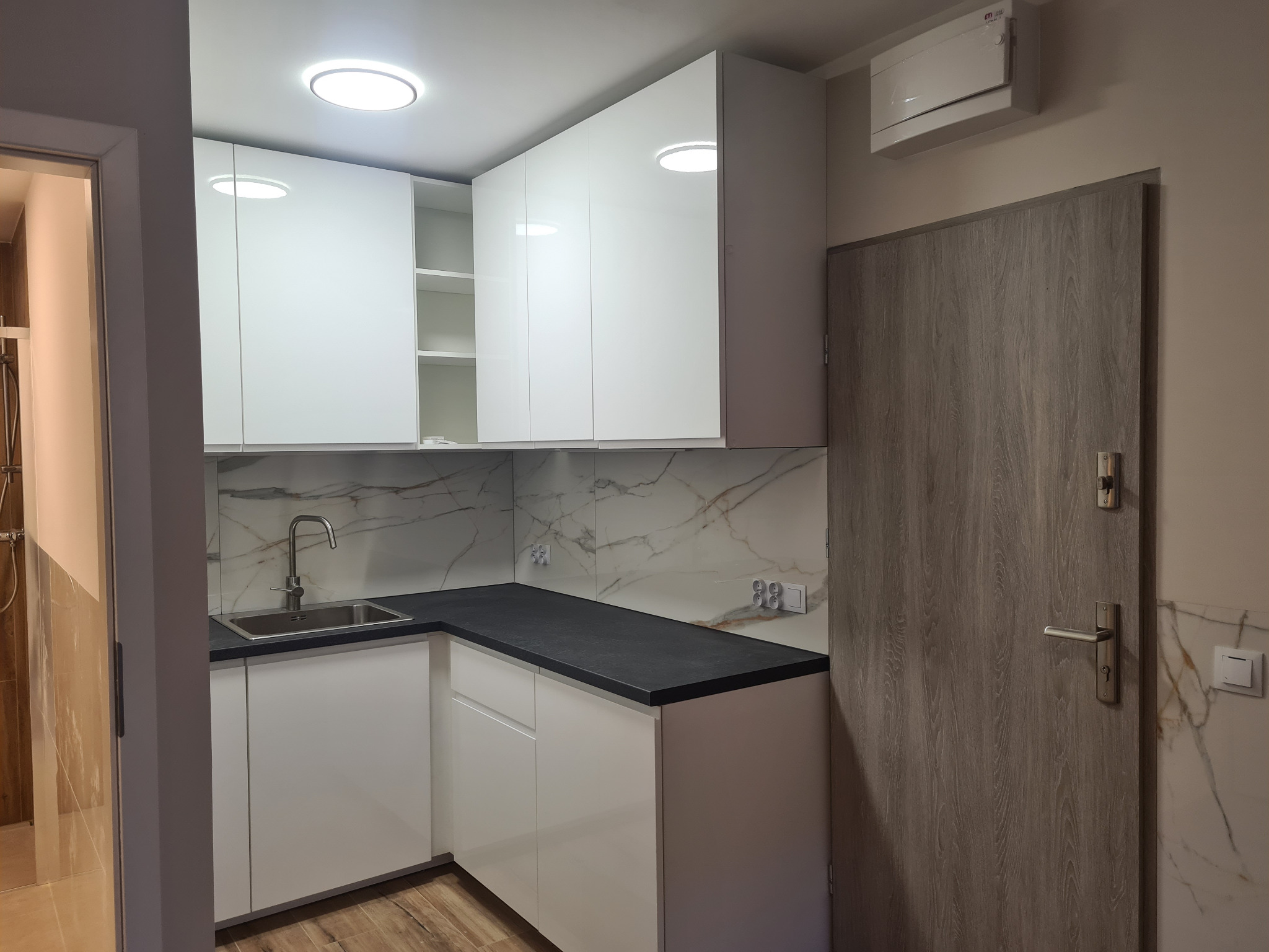Nowoczesna kuchnia z białymi, błyszczącymi szafkami, czarnym blatem i marmurowym backsplashiem. Widoczny zlew, bateria i drewnopodobna podłoga. Drzwi w odcieniach szarości.