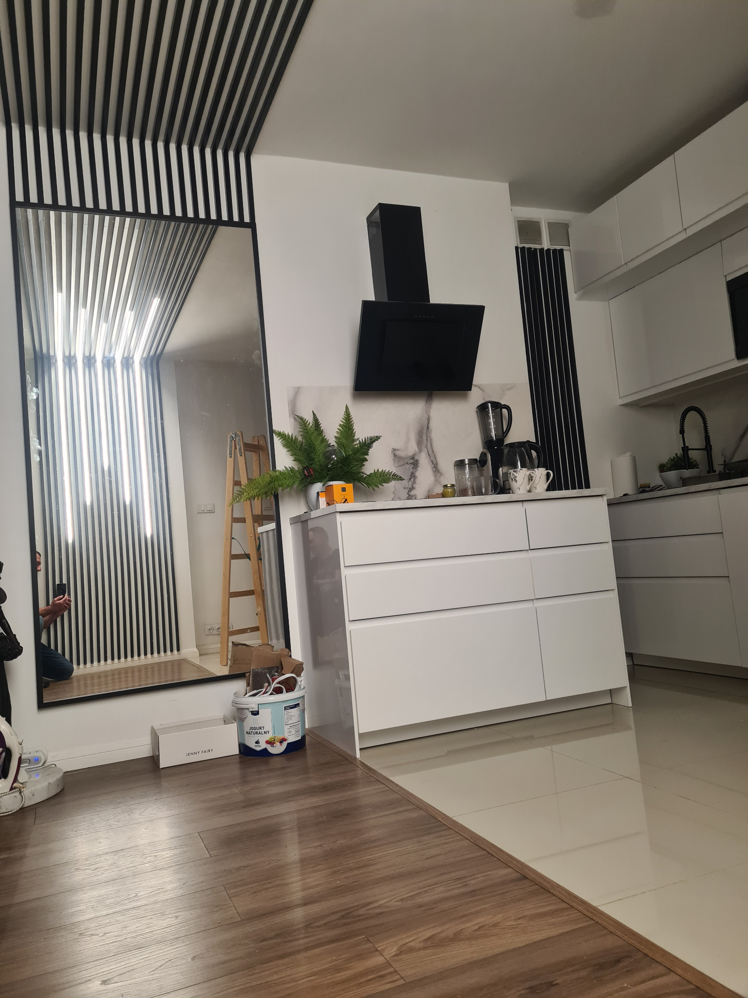 Nowoczesna kuchnia z białymi meblami, czarnym okapem i lustrem z czarnymi listwami. Na podłodze panele imitujące drewno. Widoczny fragment drabiny malarskiej.