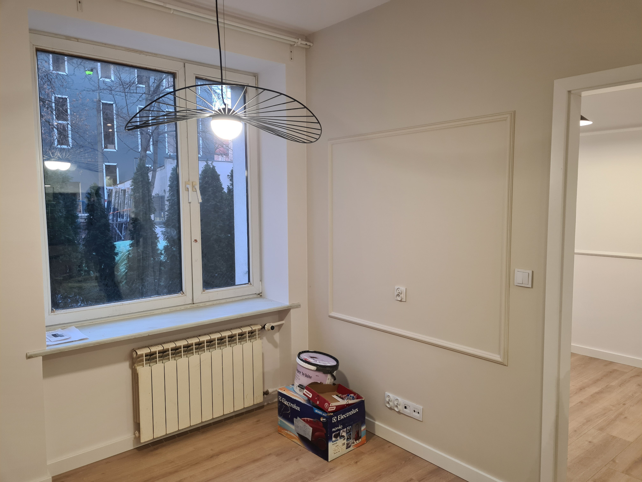 Jasne wnętrze po remoncie: kremowe ściany z dekoracyjną ramą, nowoczesna lampa, okno z widokiem na budynek. Na podłodze puszka z farbą i karton.