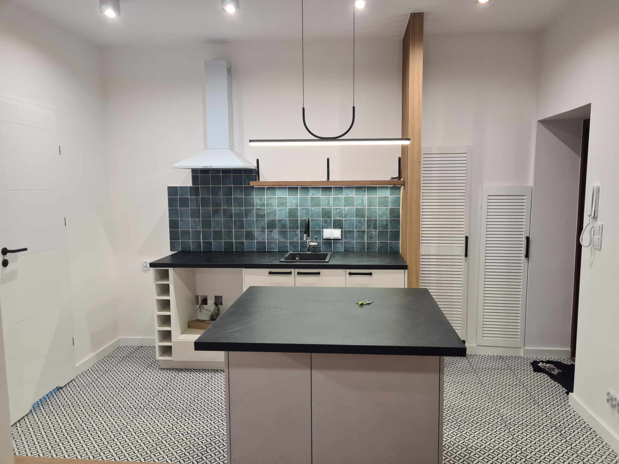 Nowoczesna kuchnia z wyspą, płytkami w odcieniach zieleni i czarnym blatem. Uwagę zwraca designerska lampa nad wyspą i wzorzysta podłoga.