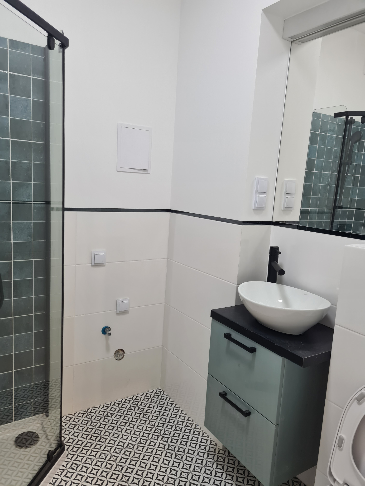 Nowoczesna łazienka po remoncie: kabina prysznicowa z ciemną glazurą, białe płytki, umywalka nablatowa na szafce w kolorze morskim i wzorzysta podłoga.