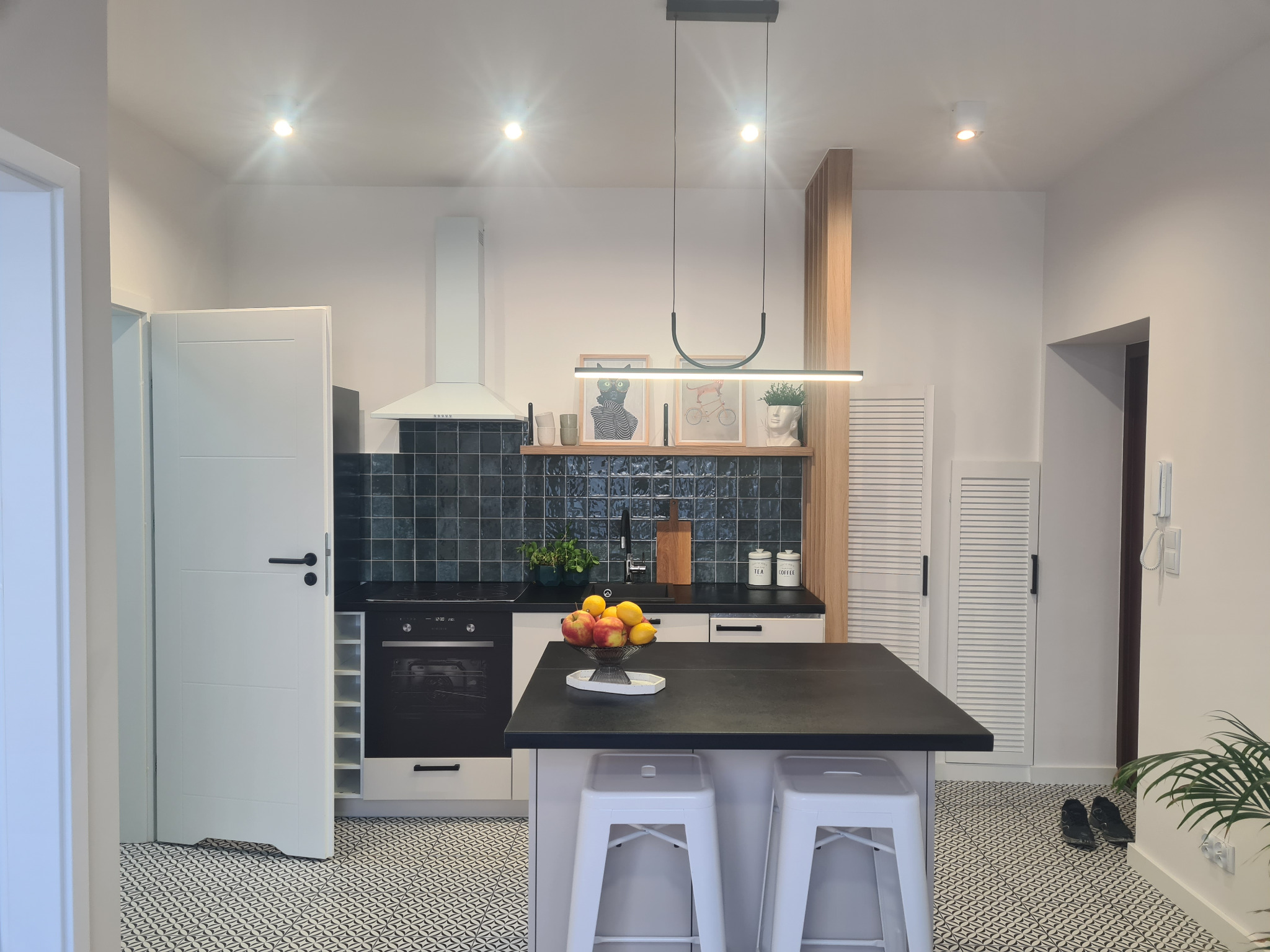 Nowoczesna kuchnia z wyspą, czarnym blatem i płytkami w odcieniach zieleni. Białe szafki, designerska lampa i wzorzysta podłoga tworzą spójną całość.