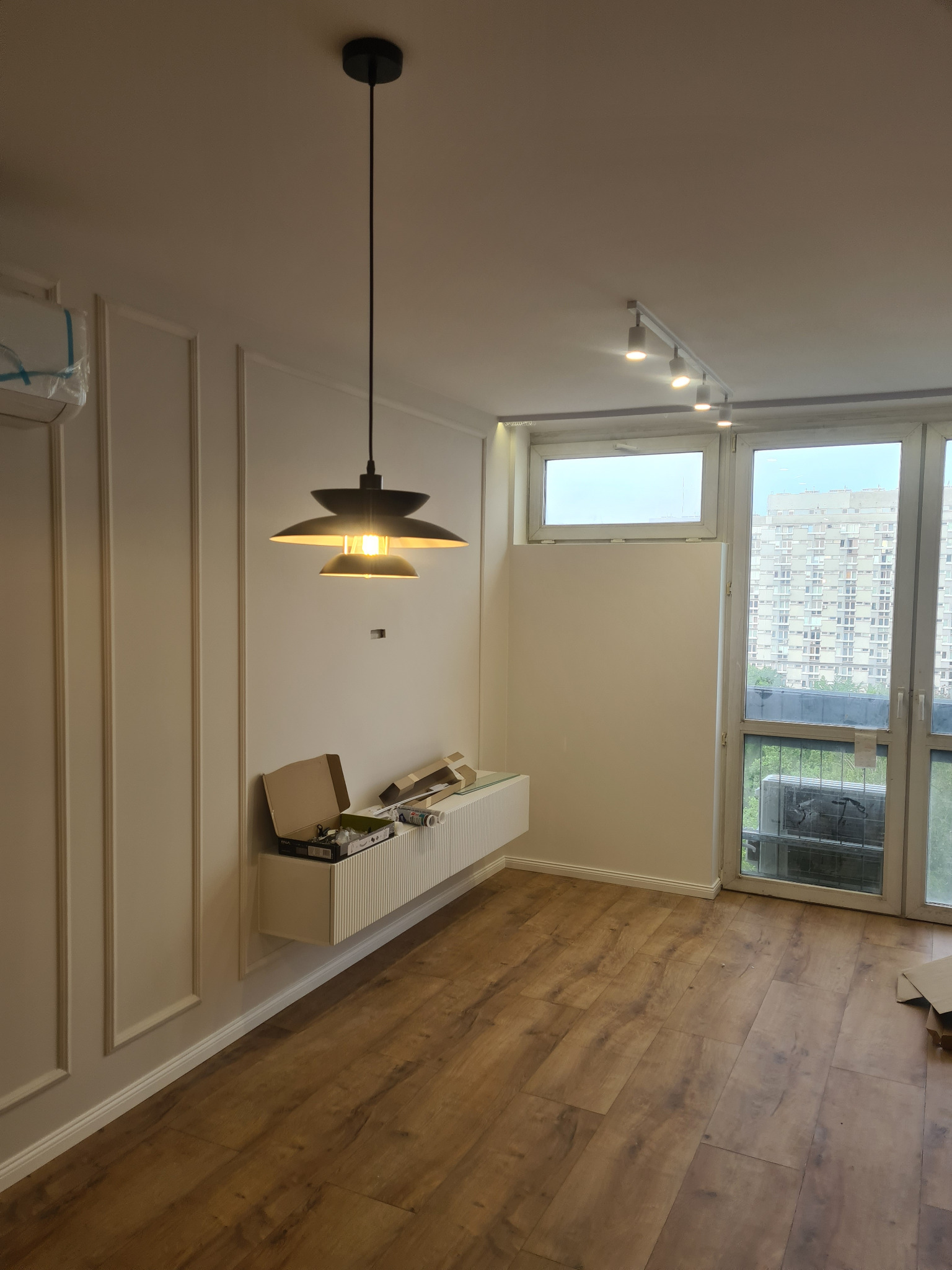 Jasne wnętrze po remoncie: panele podłogowe, białe ściany z dekoracyjnymi listwami, nowoczesne oświetlenie i wisząca szafka z akcesoriami. Widok na blok z okna.