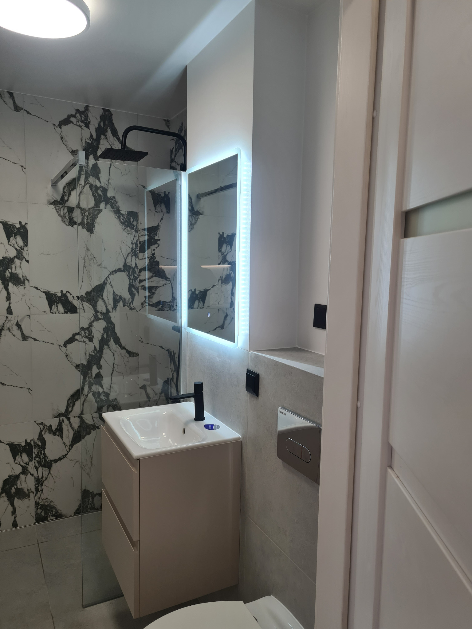 Nowoczesna łazienka po remoncie: kabina walk-in z marmurową ścianą, podświetlane lustro nad beżową szafką z umywalką, szara płytka podłogowa i białe drzwi.